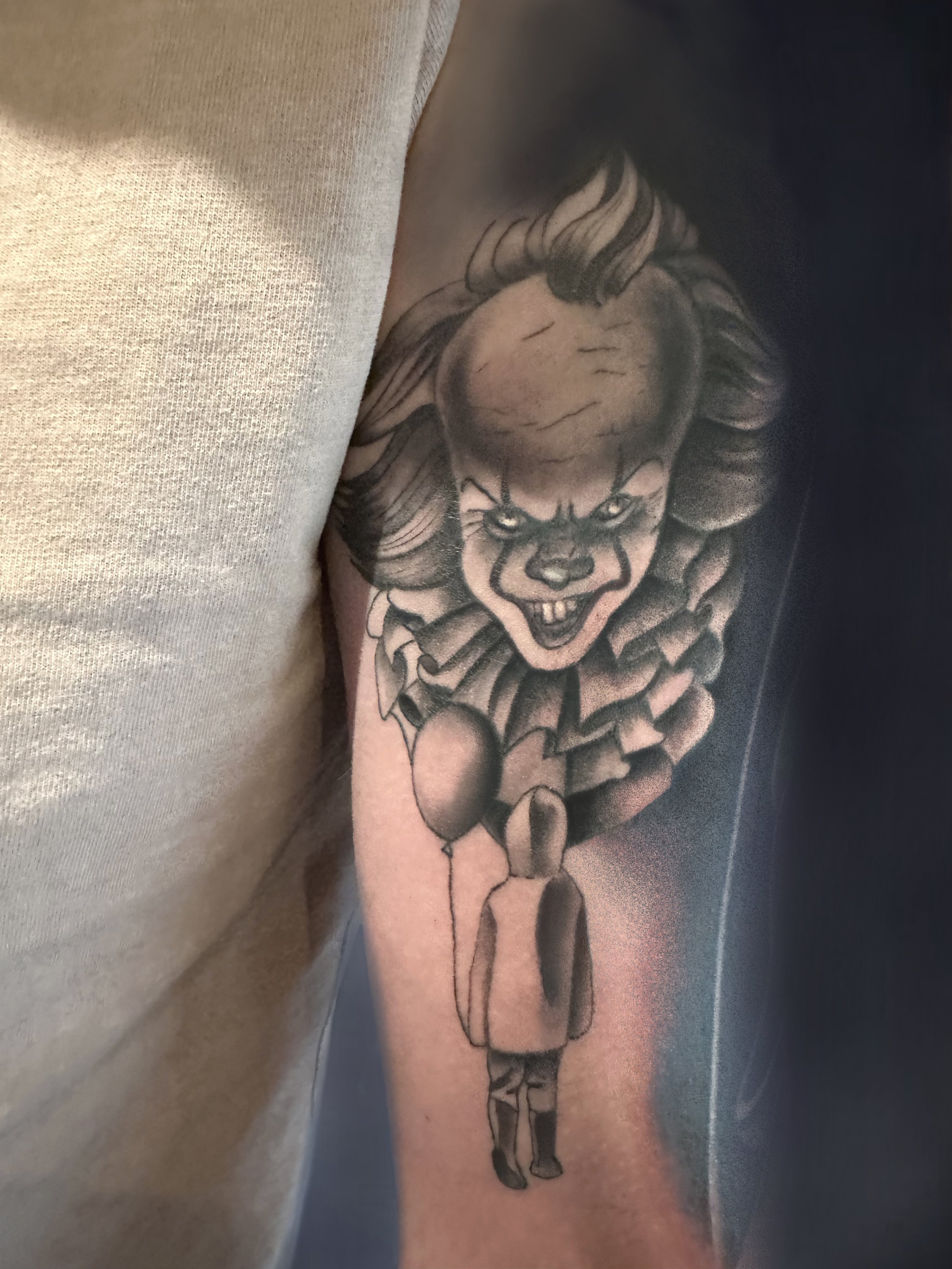 Tattoo of a clown face with sharp features and a sinister smile on a person's upper arm, with a figure holding balloons underneath.
