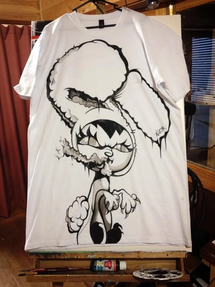 A white T-shirt hanging on a hook. The shirt has a black and white cartoon-like graphic of a character with large ears, a wide-open mouth, and a cigar in its mouth, smoking smoke. The design is bold and stylized.