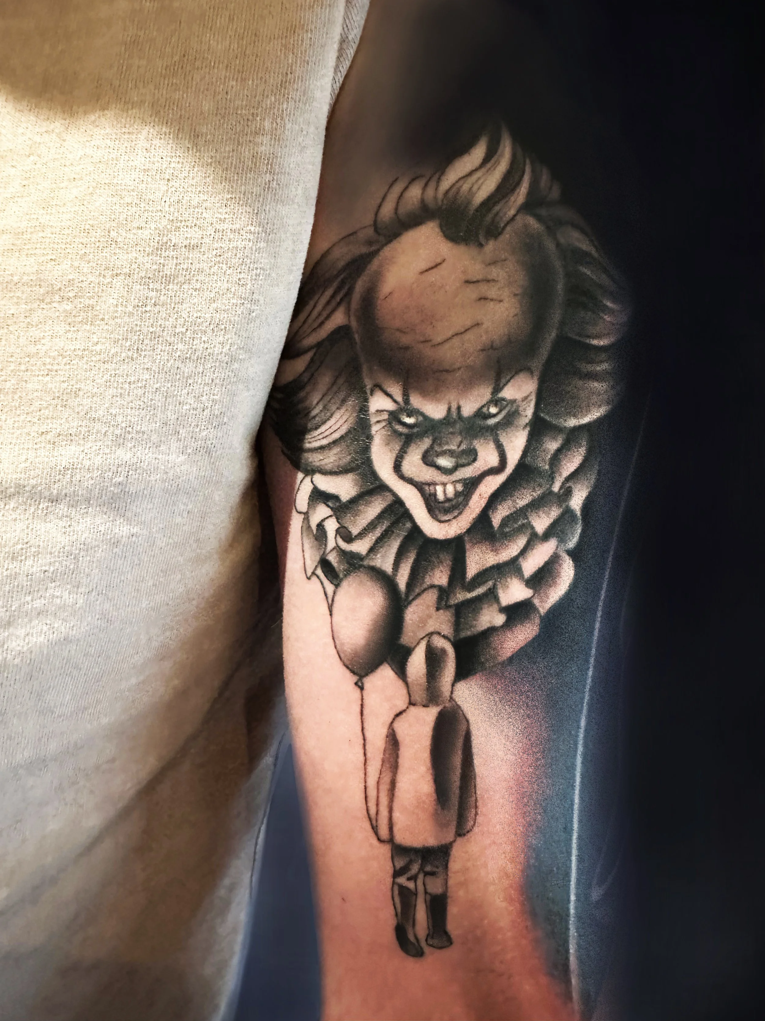 Tattoo of a sinister clown face with a large head and clown makeup, appearing to float above a figure of a girl holding a balloon.