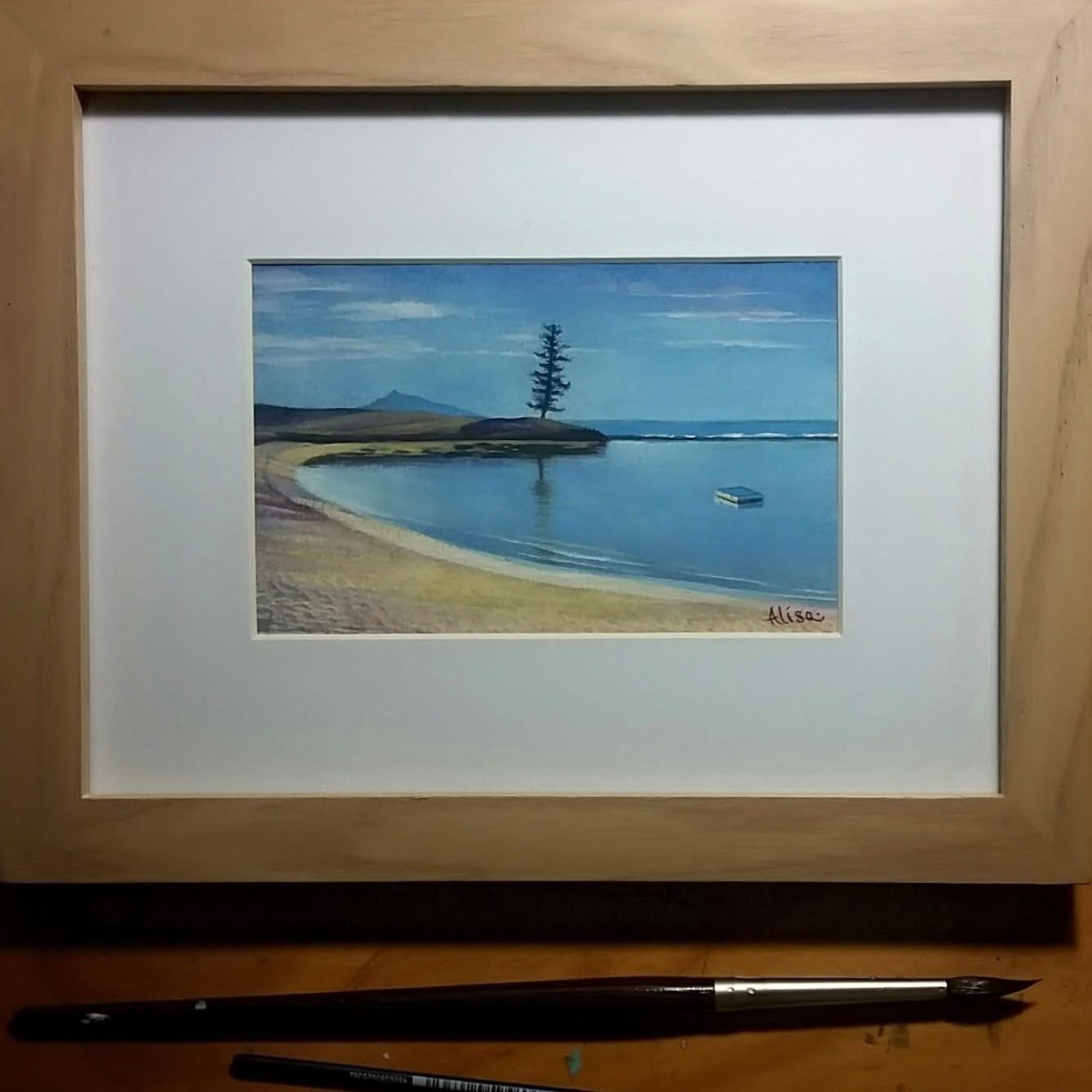 A framed painting of a peaceful lakeside scene with a beach, calm water, a tall tree on a small island, and mountains in the background, signed by Alisa.