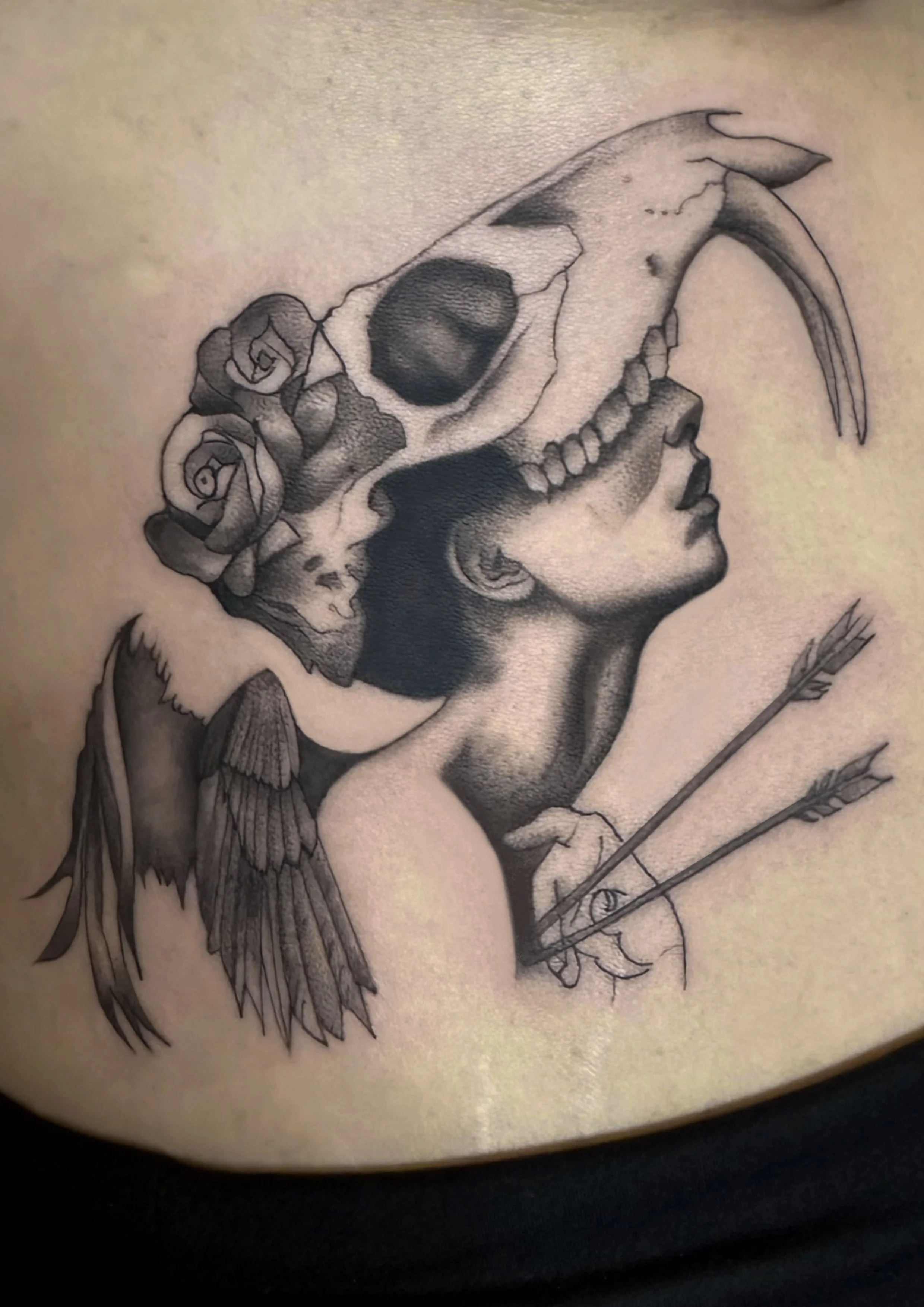 Tattoo of a woman wearing an animal skull with horns on her head, flowers, bird wings, and arrows.