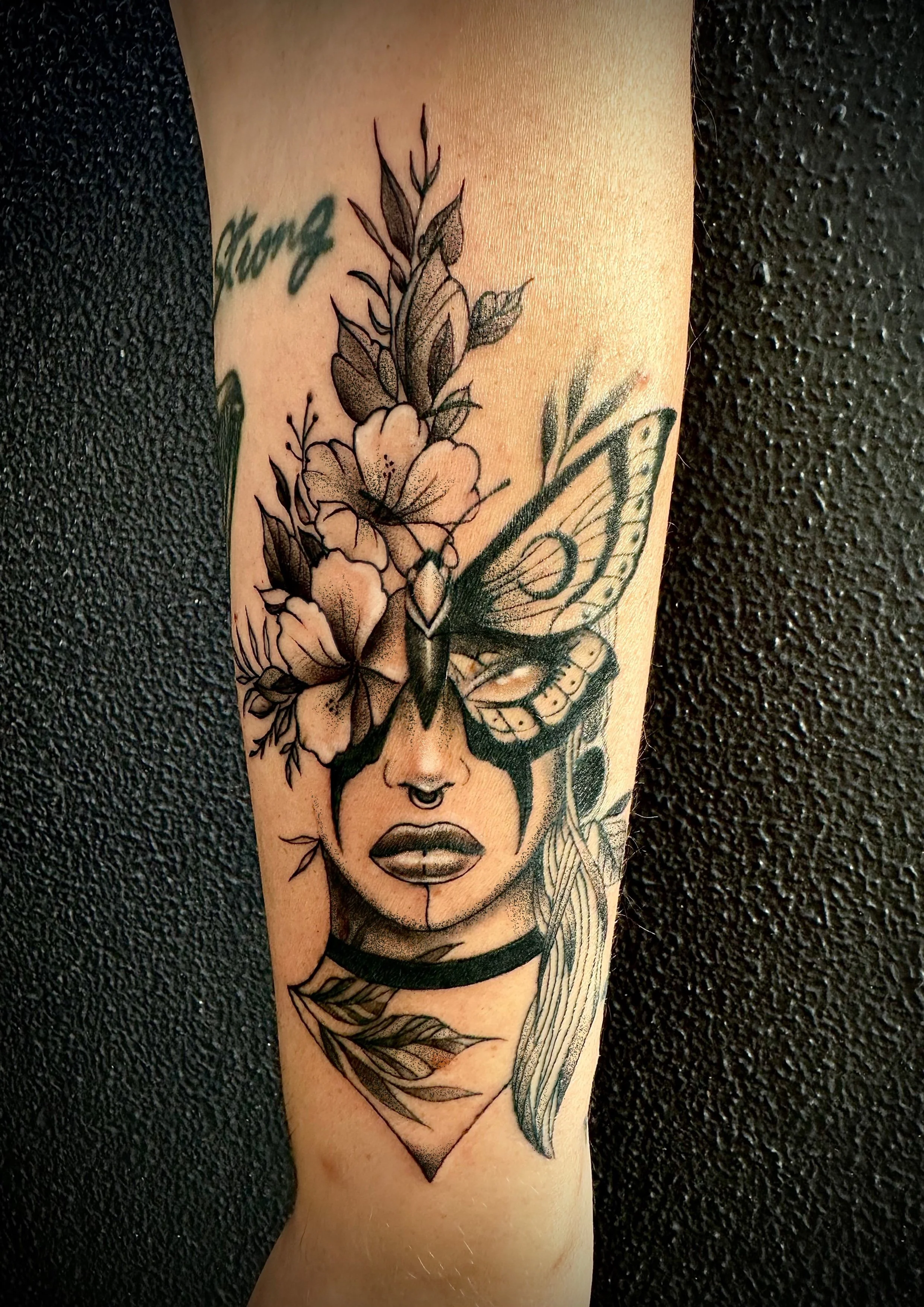 A black and gray tattoo depicting a woman's face with closed eyes and bold lips, with a butterfly and flowers growing from her forehead, and a leaf below her chin.