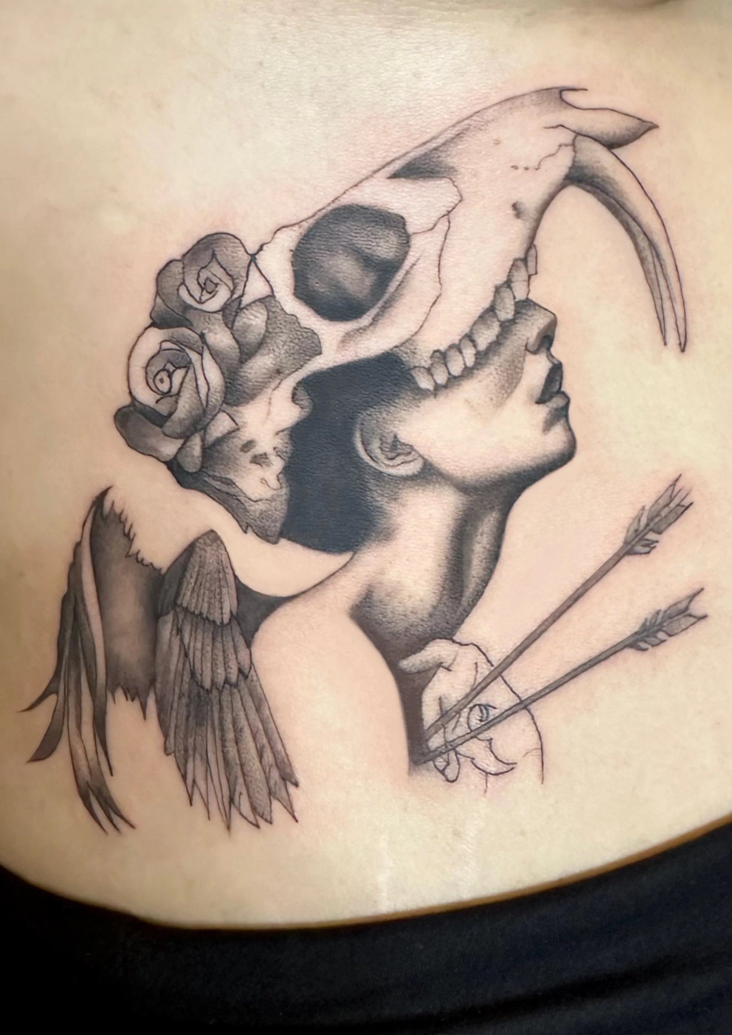 Tattoo of a woman's profile with a skull, roses, angel wings, and arrows.