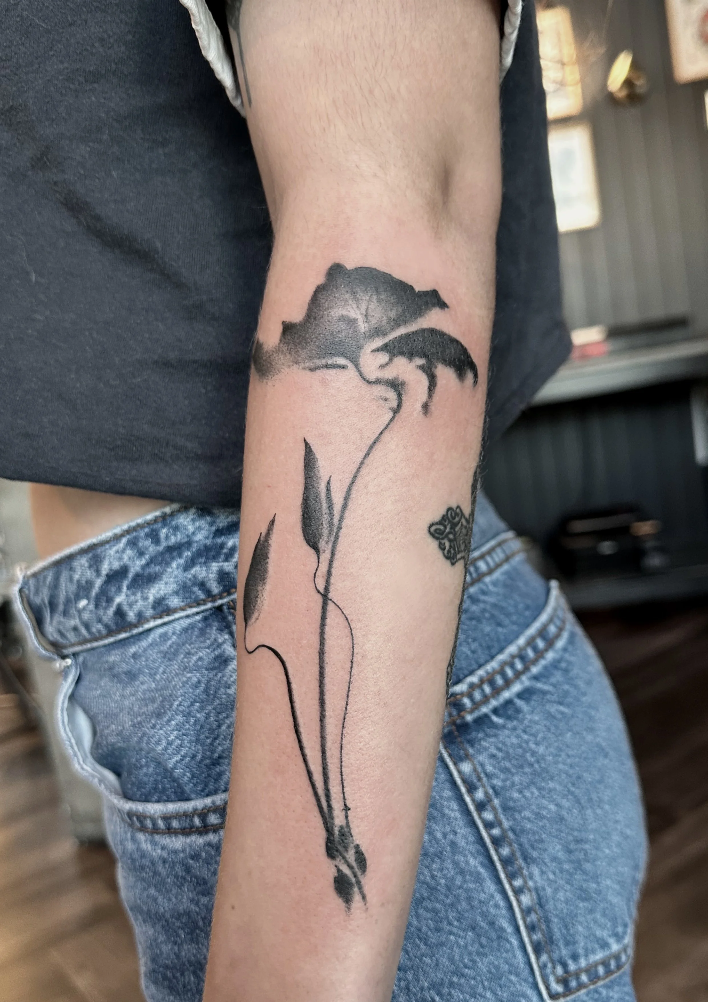 Black silhouette tattoo of a flower with a stem, leaves, and a butterfly on the person's arm.