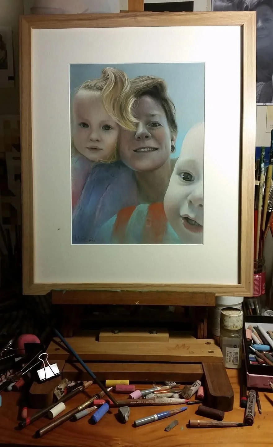 Framed watercolor portrait of a woman and two children. The woman has short hair and is smiling. The girl on the left has blonde hair, and the baby on the right has light hair with a curious expression. The artwork is set on a studio table with drawi