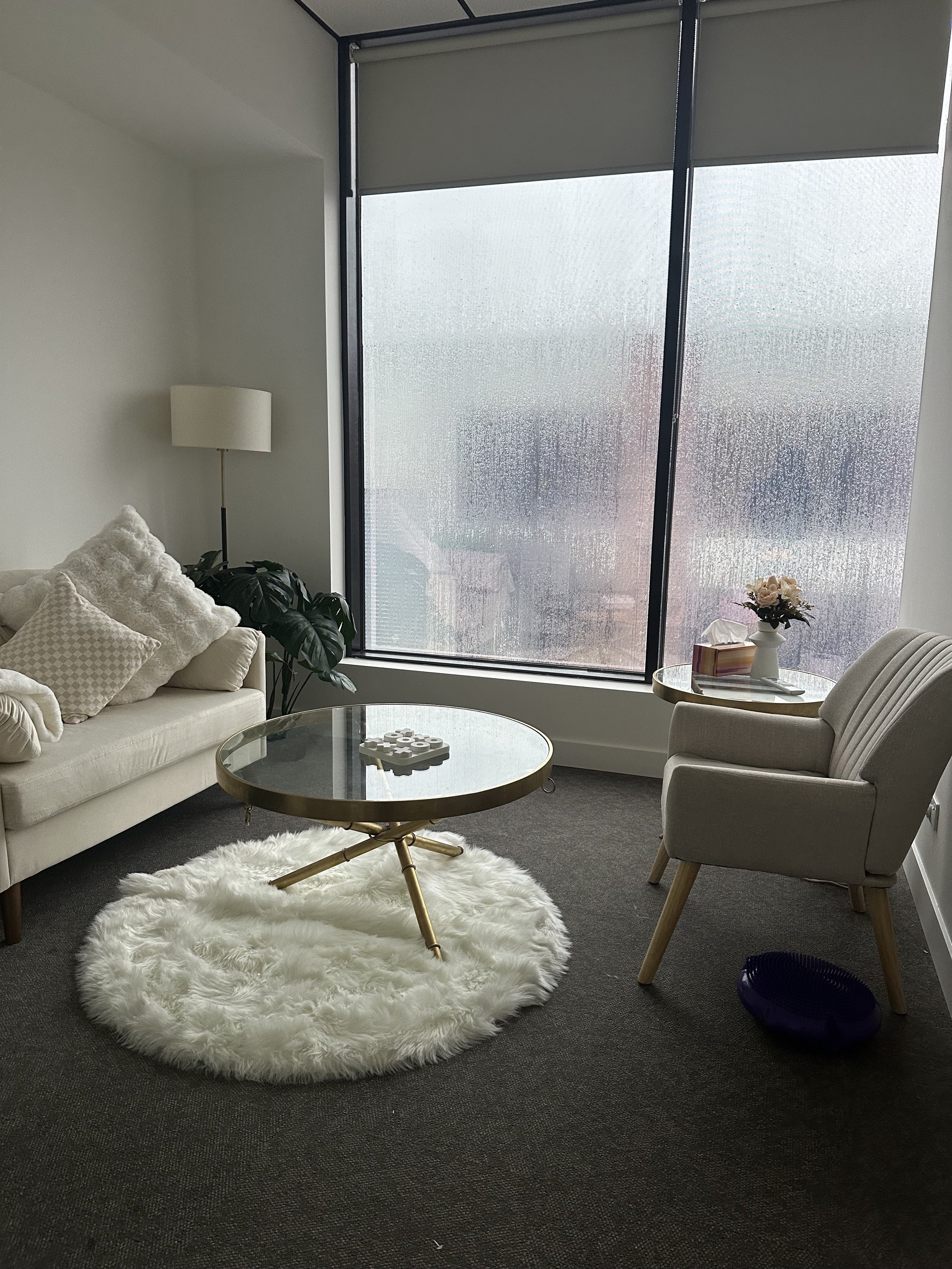 A living room corner with a white sofa, plush pillows, a tall floor lamp, a large window with frosted glass, a side table with a flower arrangement and books, a beige armchair, a round glass coffee table on a fluffy white rug, and a dark blue object on the floor.