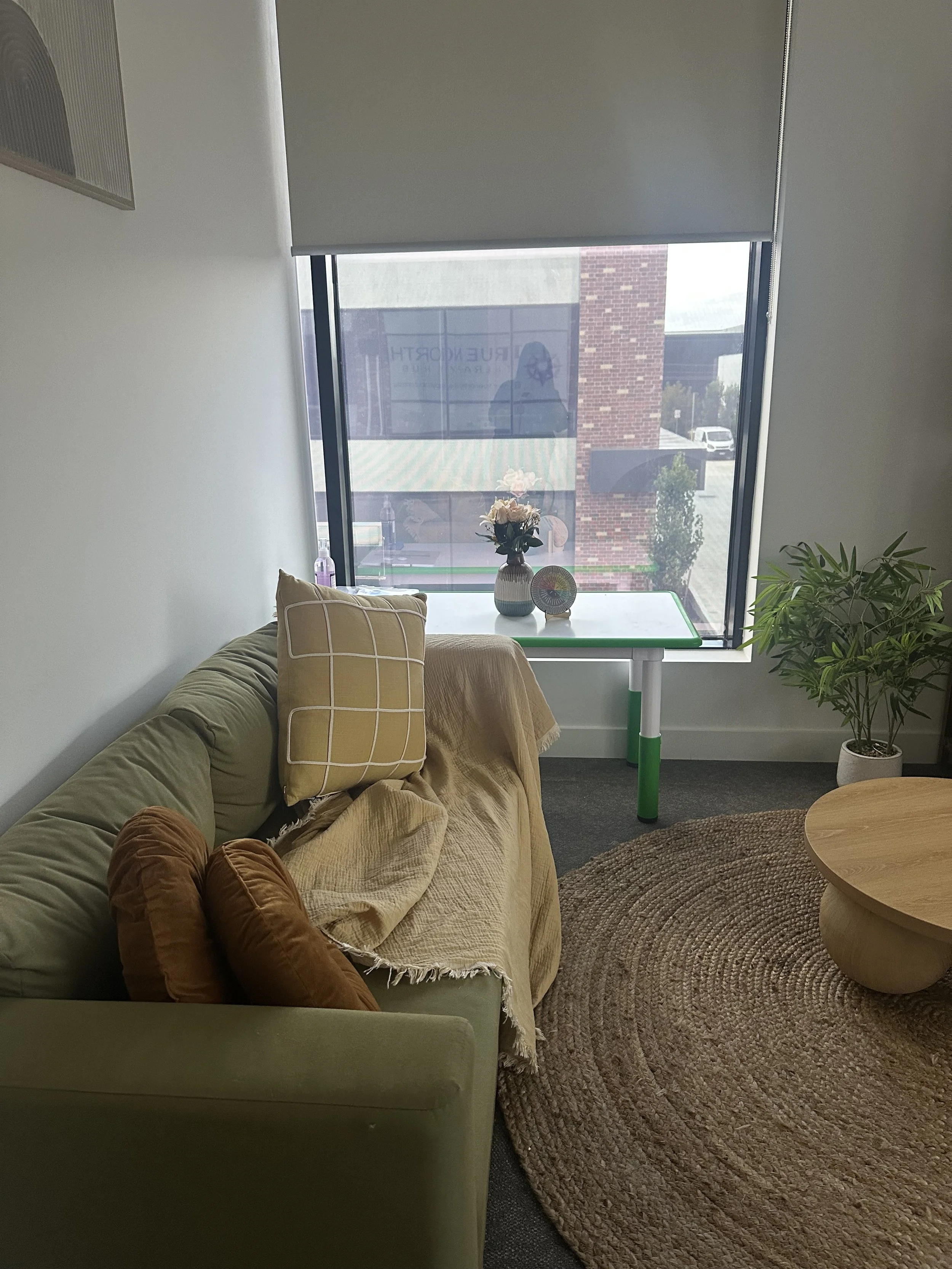 Interior of a cozy living room with a green sofa, a beige blanket, brown and beige cushions, a round woven rug, a small round wooden coffee table, a window with a view of brick buildings outside, and a white table with vases of flowers and decorative items.