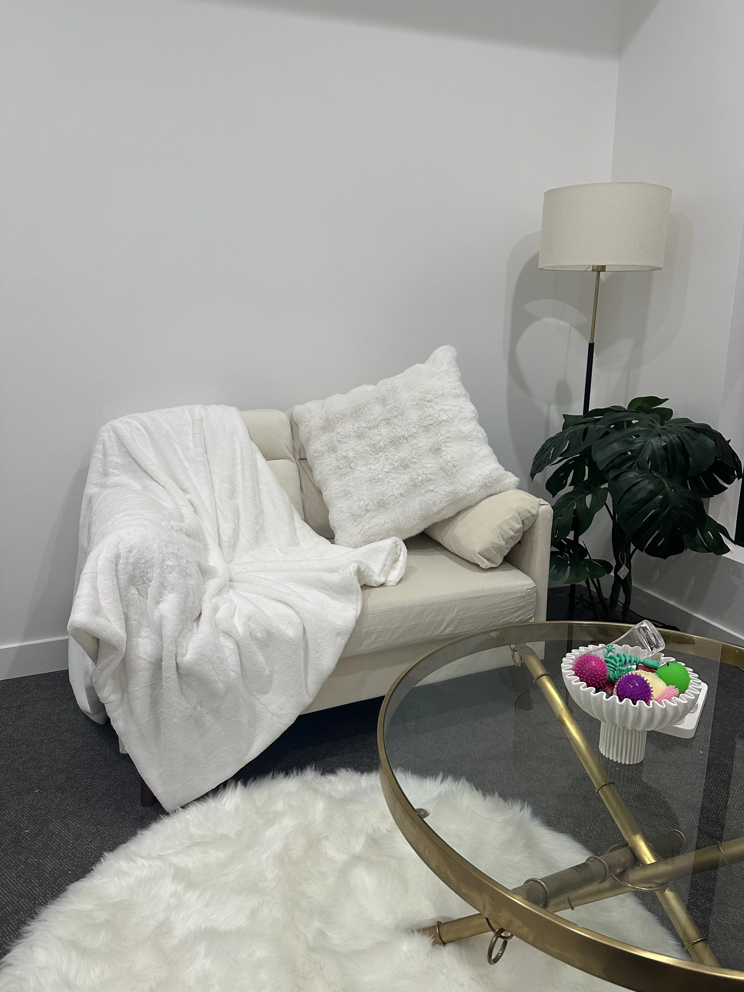 A cozy living room corner with a white sofa draped with a white blanket, a fluffy white pillow, and a beige pillow. There is a tall floor lamp with a beige lampshade next to a large green leafy plant. A round glass-top coffee table with a gold frame holds a white decorative bowl filled with colorful festive ornaments. A white shag rug covers part of the dark carpet floor.