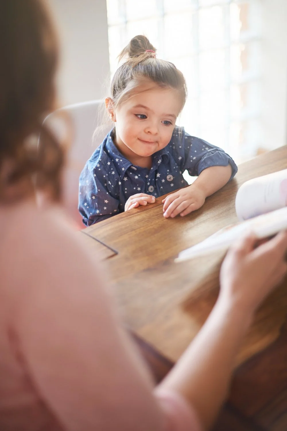 A young girl with a bun hairstyle wearing a blue star-patterned shirt, sitting at a wooden table, looking at a book held by an adult.