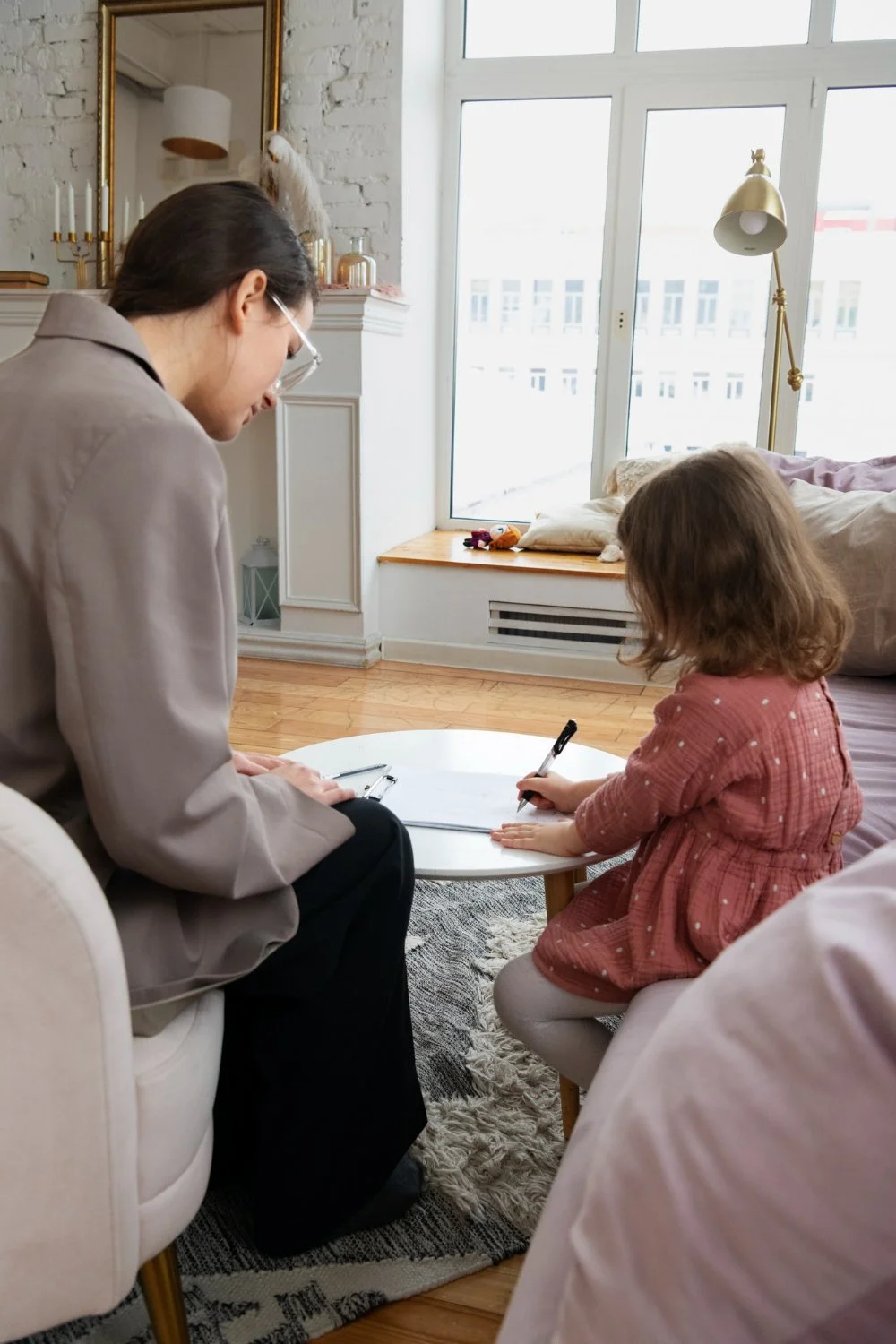 A woman and a young girl are seated on sofas around a small white coffee table, with the girl writing in a notebook while the woman observes. The room has a large window, a lamp, and decorative items on the mantel.