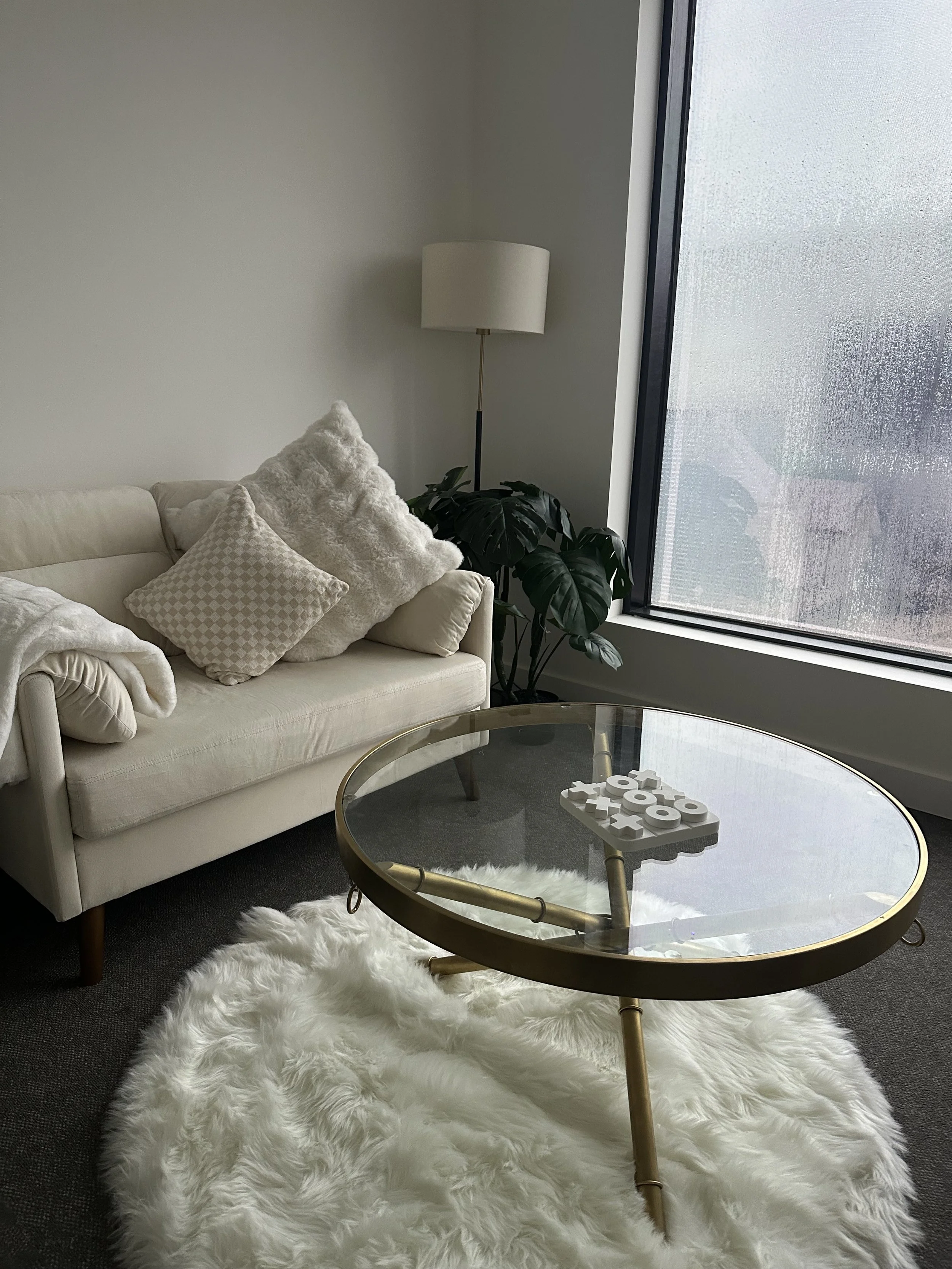 Living room with a white sofa, various pillows, a tall floor lamp, a large window with condensation, a potted plant, a glass coffee table with a white decorative tic-tac-toe game, and a white fluffy rug.