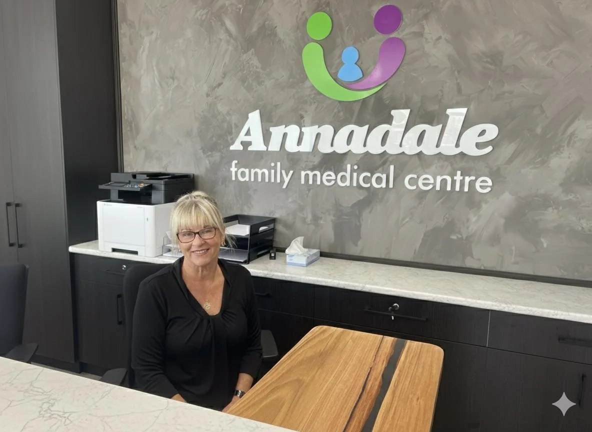 A woman sitting at a reception desk in the Annadale Family Medical Centre, smiling and wearing glasses and a black top.