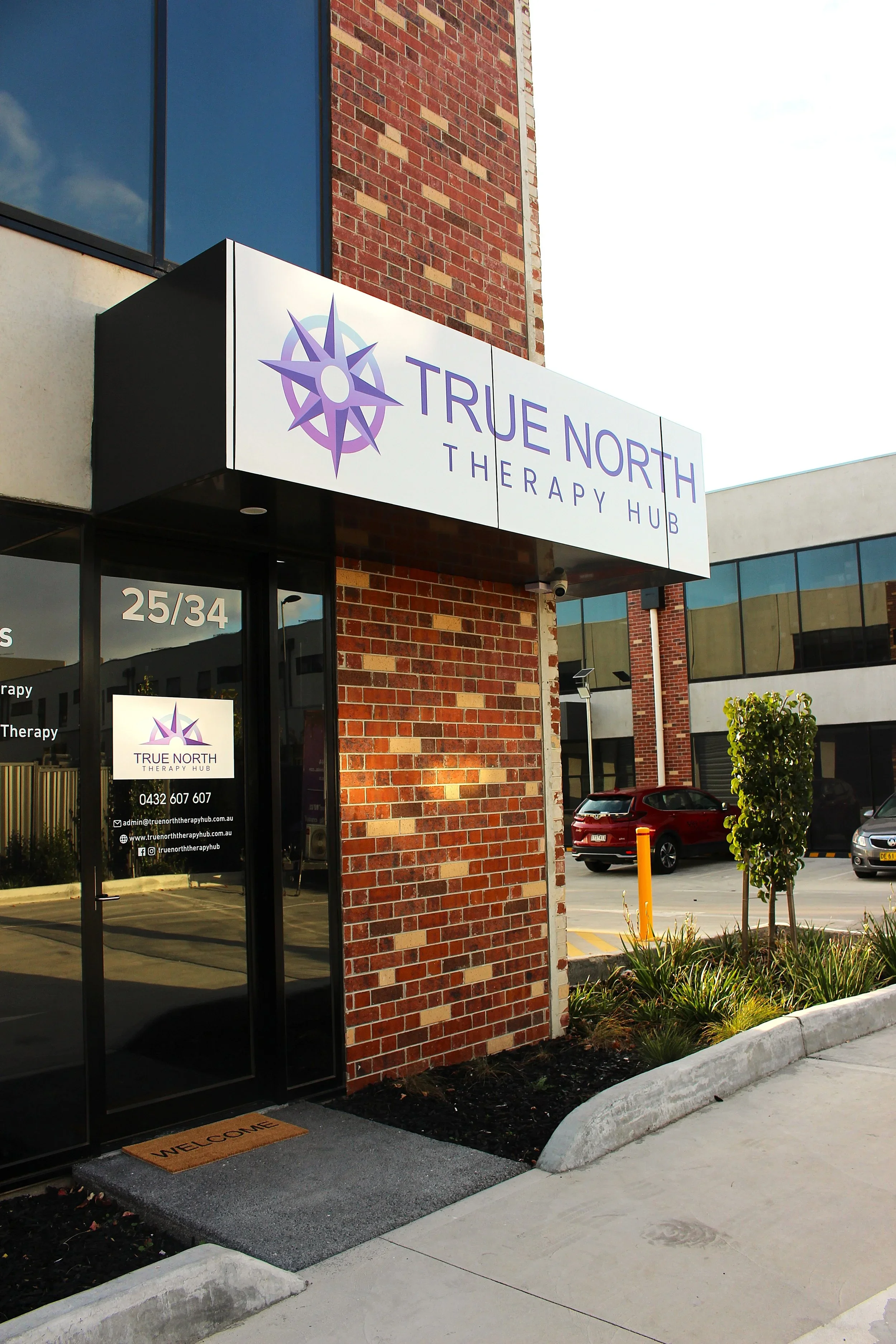 Exterior view of a therapy hub with a sign reading 'True North Therapy Hub' in purple and white, featuring a purple compass logo. The entrance has a glass door with contact information and a welcome mat.