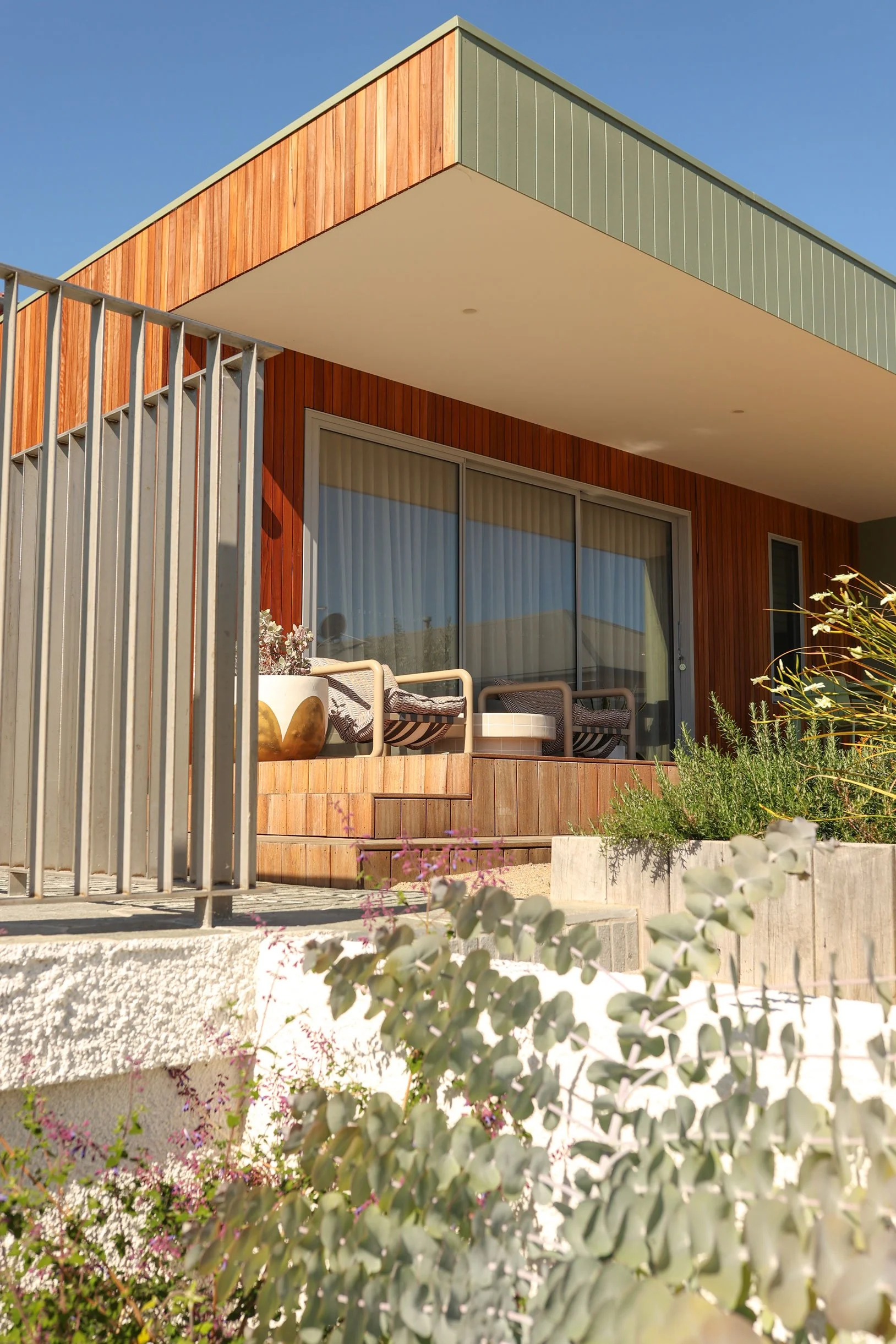 Modern house with wooden and green siding, large sliding glass doors, outdoor patio with chairs and plants, and a garden in foreground.