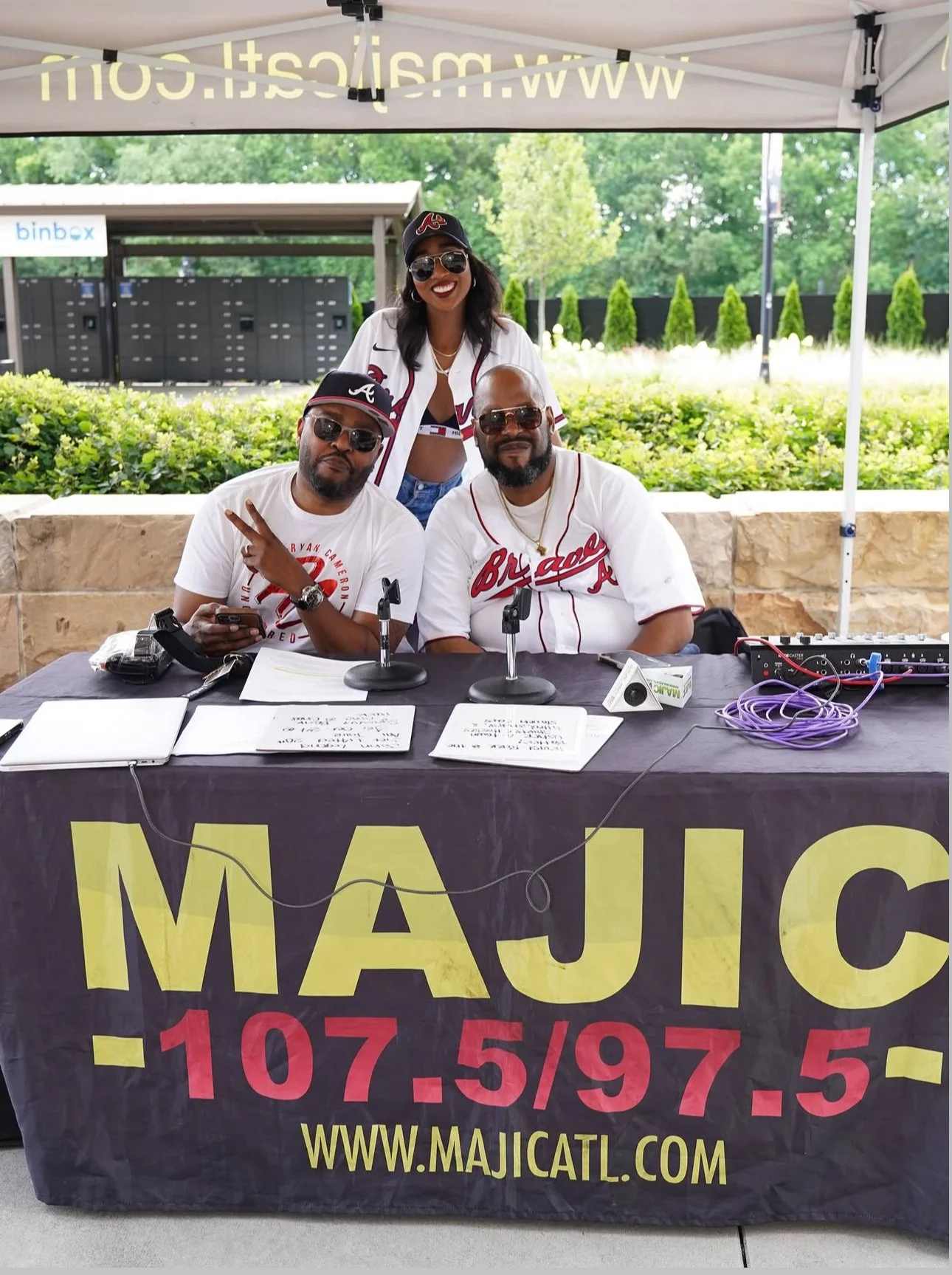 Three people sitting at a table under a canopy, with a woman standing behind two men. They are wearing Atlanta Braves baseball jerseys and hats, all wearing sunglasses. The table has microphones, documents, and electronic equipment. The backdrop shows greenery and a public space.