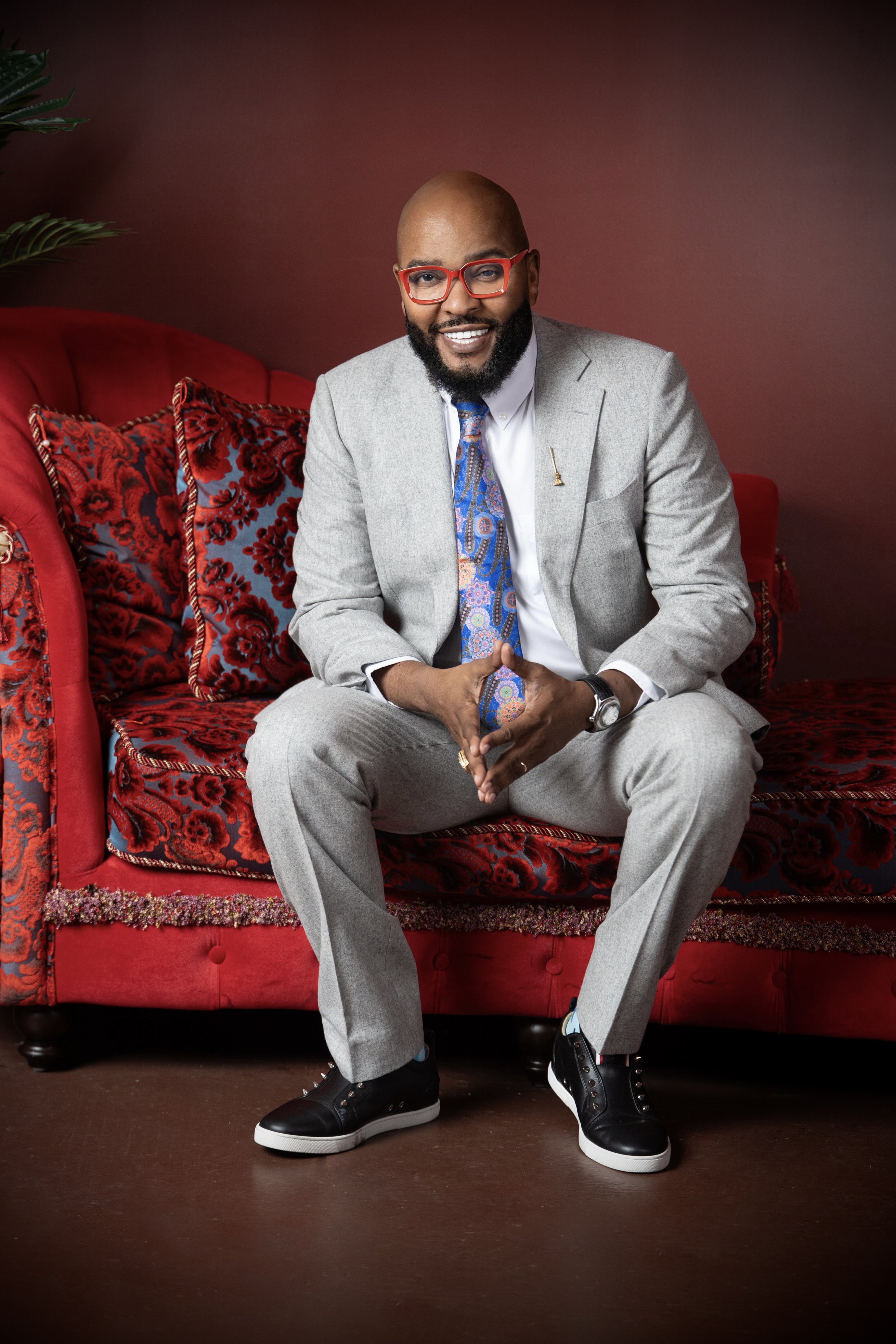A smiling man with a beard and bald head wearing red glasses, a light gray suit, a colorful patterned tie, and black sneakers. He is sitting on a red velvet sofa with ornate floral pillows and a dark background.