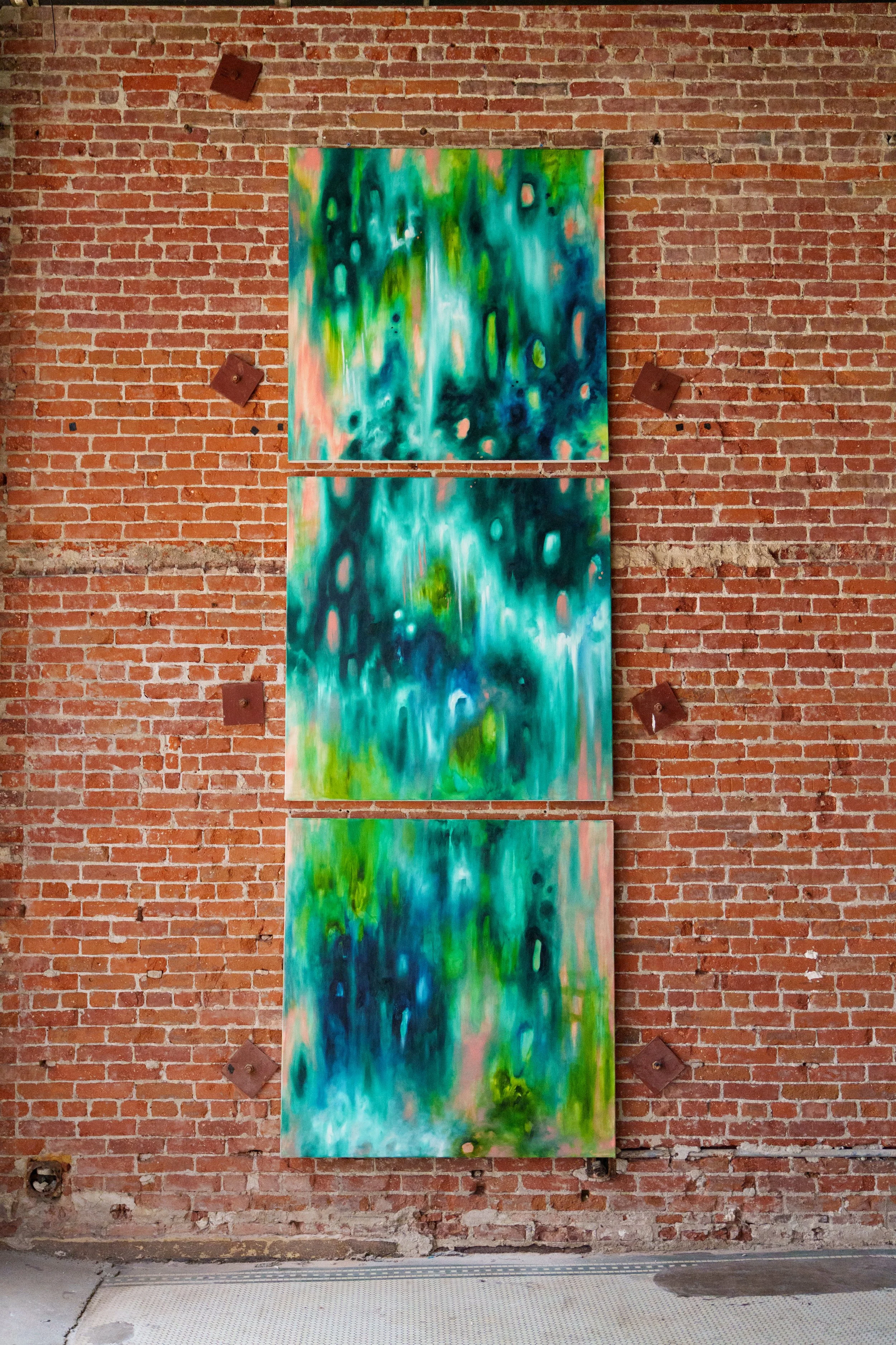 Three abstract jungle paintings with green and pink hues hanging on a brick wall, secured with metal brackets.