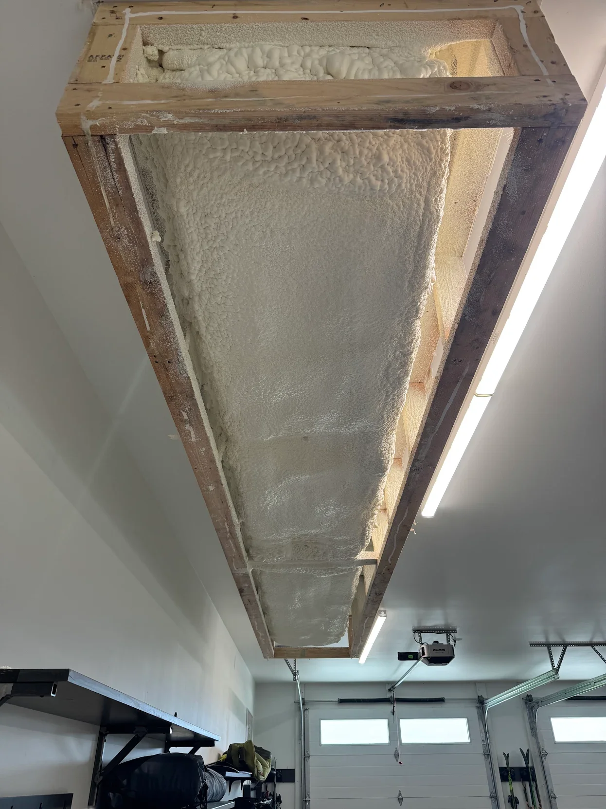 Ceiling insulation installation in a garage, showing Base Camp Spray Foam insulation inside a wooden frame.