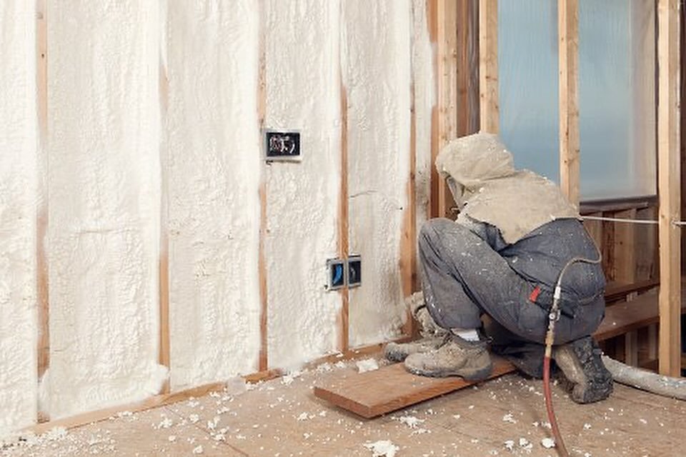 Base Camp Spray Foam, Worker crouched down, applying spray foam insulation to a wall with exposed wooden studs during construction or renovation. Flathead Valley, Montana