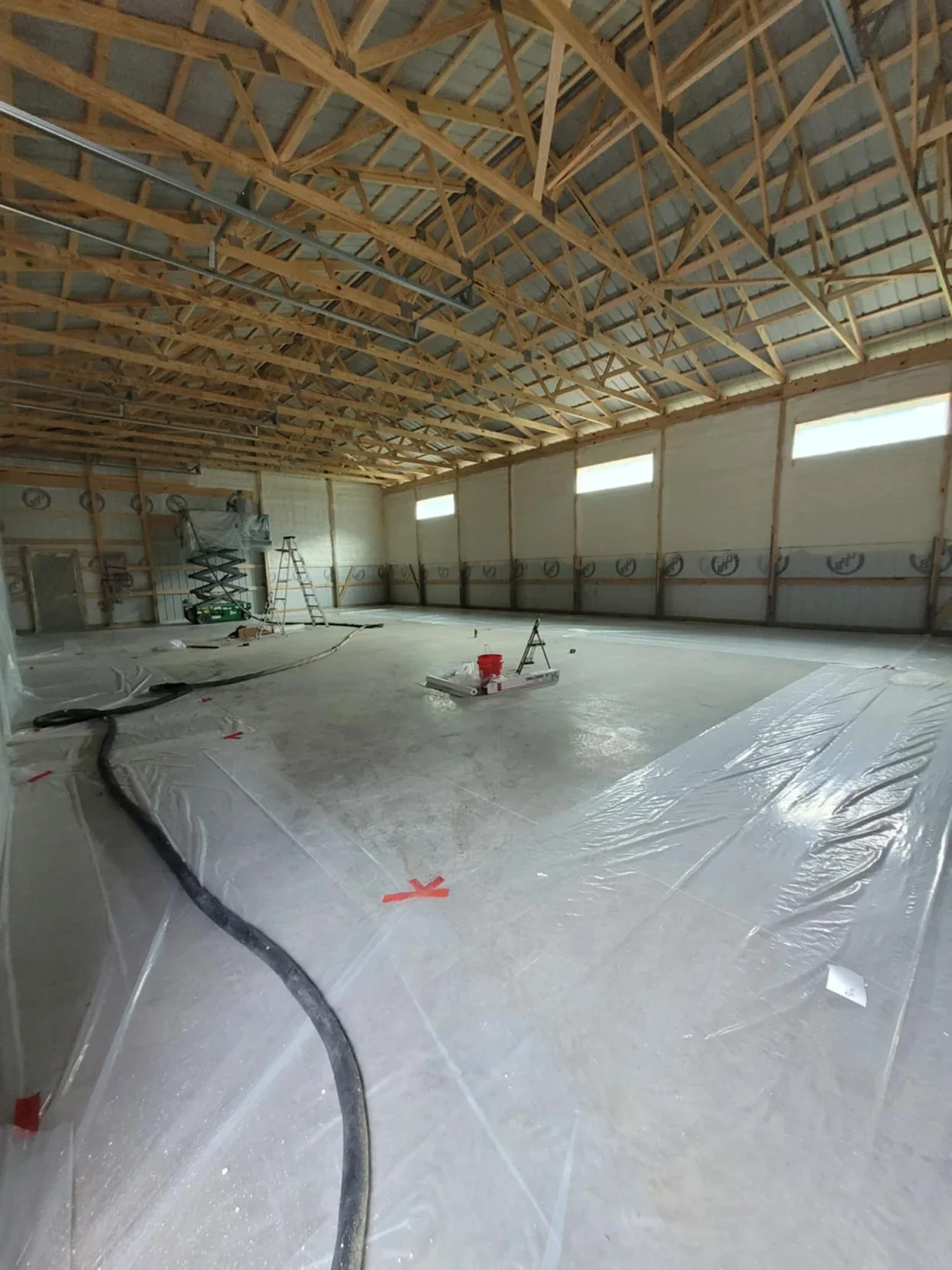 Interior of a building under construction with exposed wooden roof trusses, plastic sheeting on the floor, and construction tools and equipment including ladders, a bucket, and a scissor lift. Base Camp Spray Foam