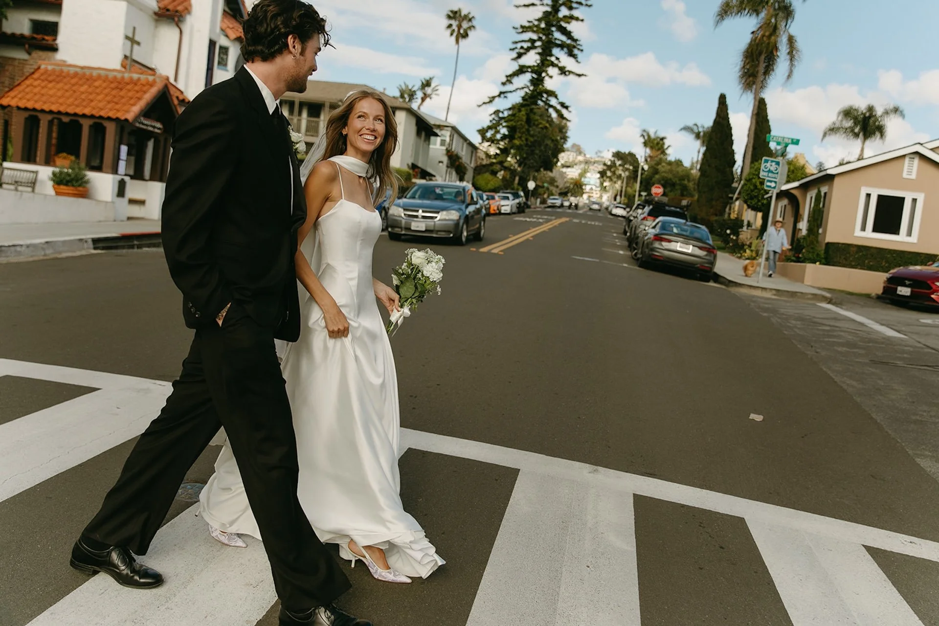 Newly married couple walking across a crosswalk together outside a Spanish-style church during their Laguna Beach wedding portraits.