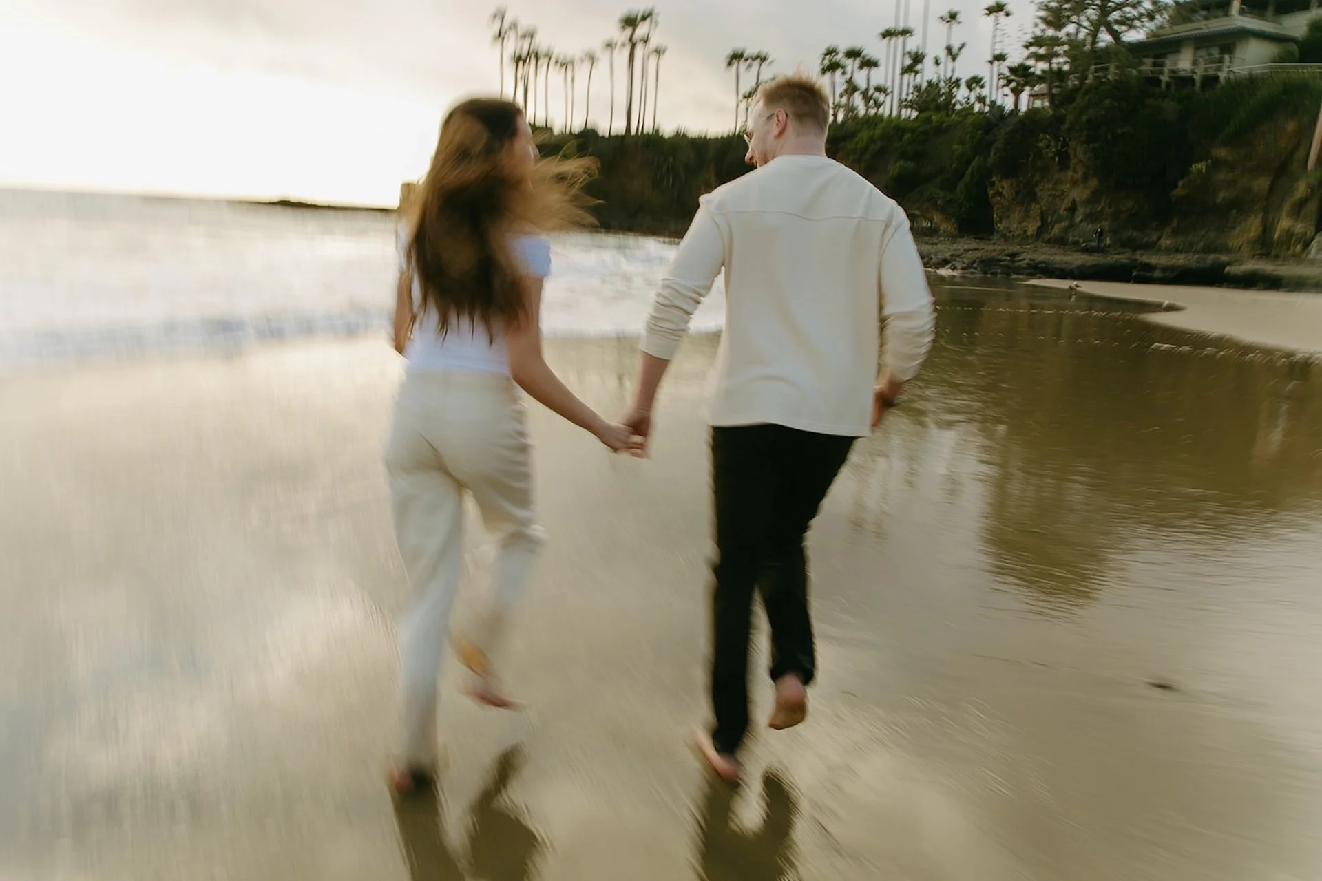 Couple running hand in hand along the shoreline, barefoot on wet sand, motion blur emphasizing movement and joy during a candid beach engagement session.
