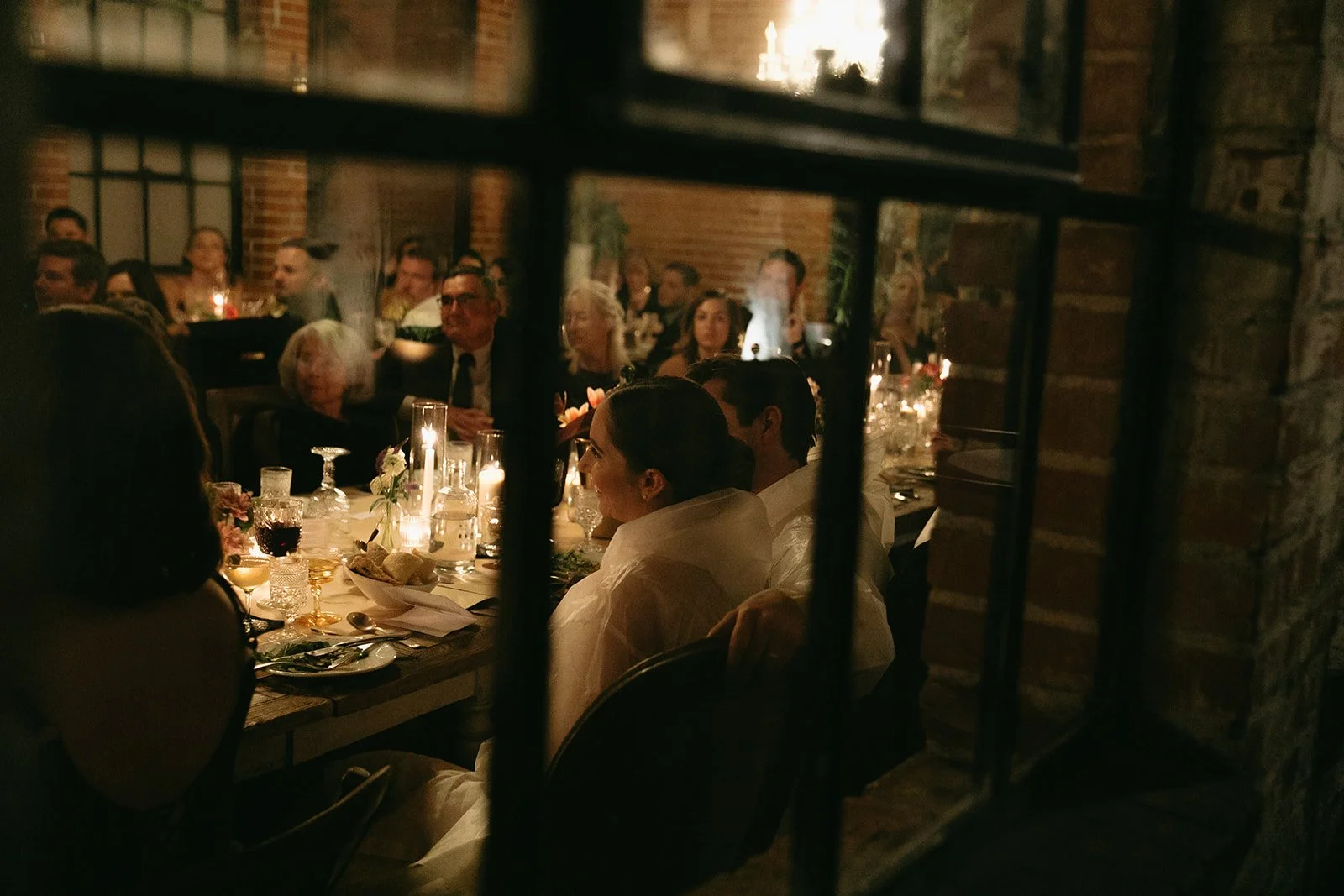 A candid moment of the bride and groom sitting at their wedding reception table and listening to their wedding guest speeches through a window pane.