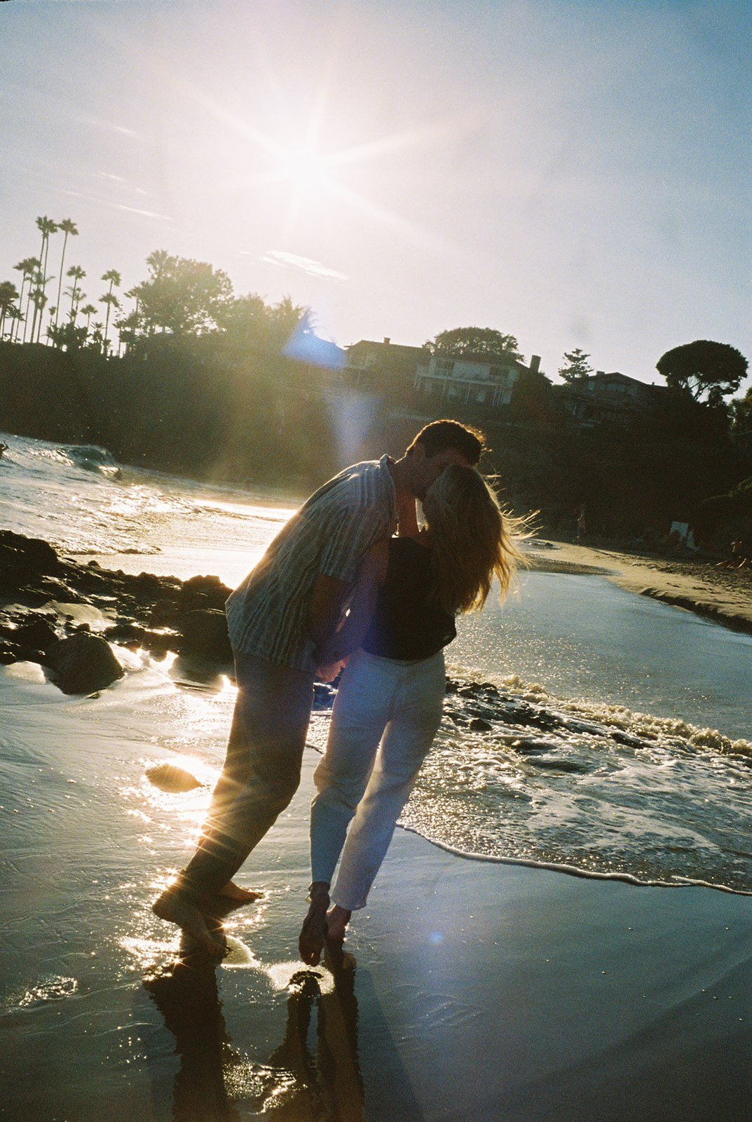 Engaged couple sharing a kiss on the beach with the sun flaring in the background—perfect moment captured with engagement photography tips.