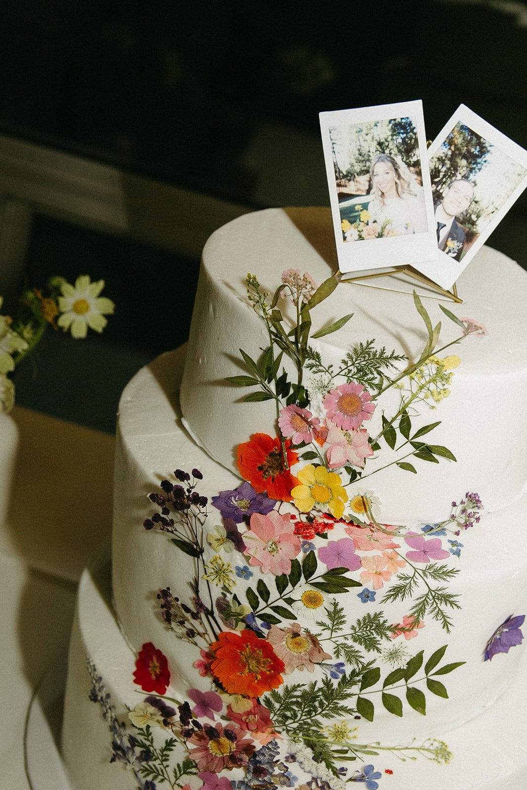 Wedding cake decorated with edible flowers and polaroid pictures of the couple, capturing a moment of their celebration for a unique wedding idea.