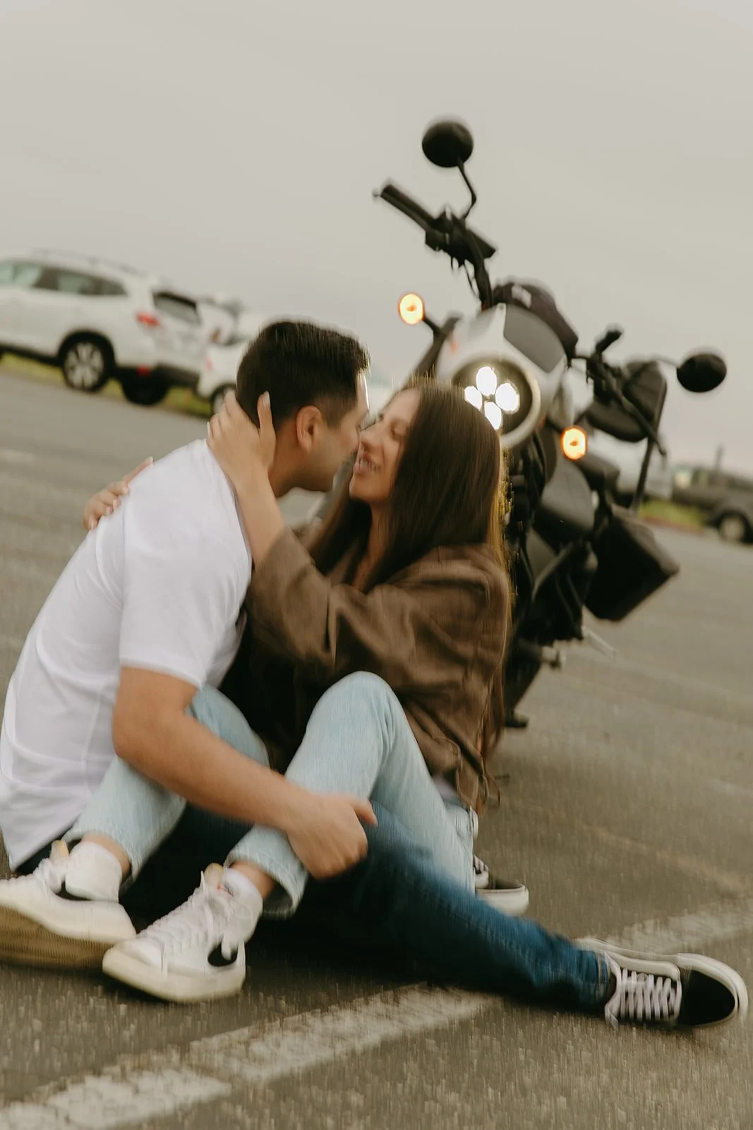A cozy moment captured as a couple sits in a parking lot, leaning into each other with a motorcycle in the background, a fun take on romantic engagement photo ideas.