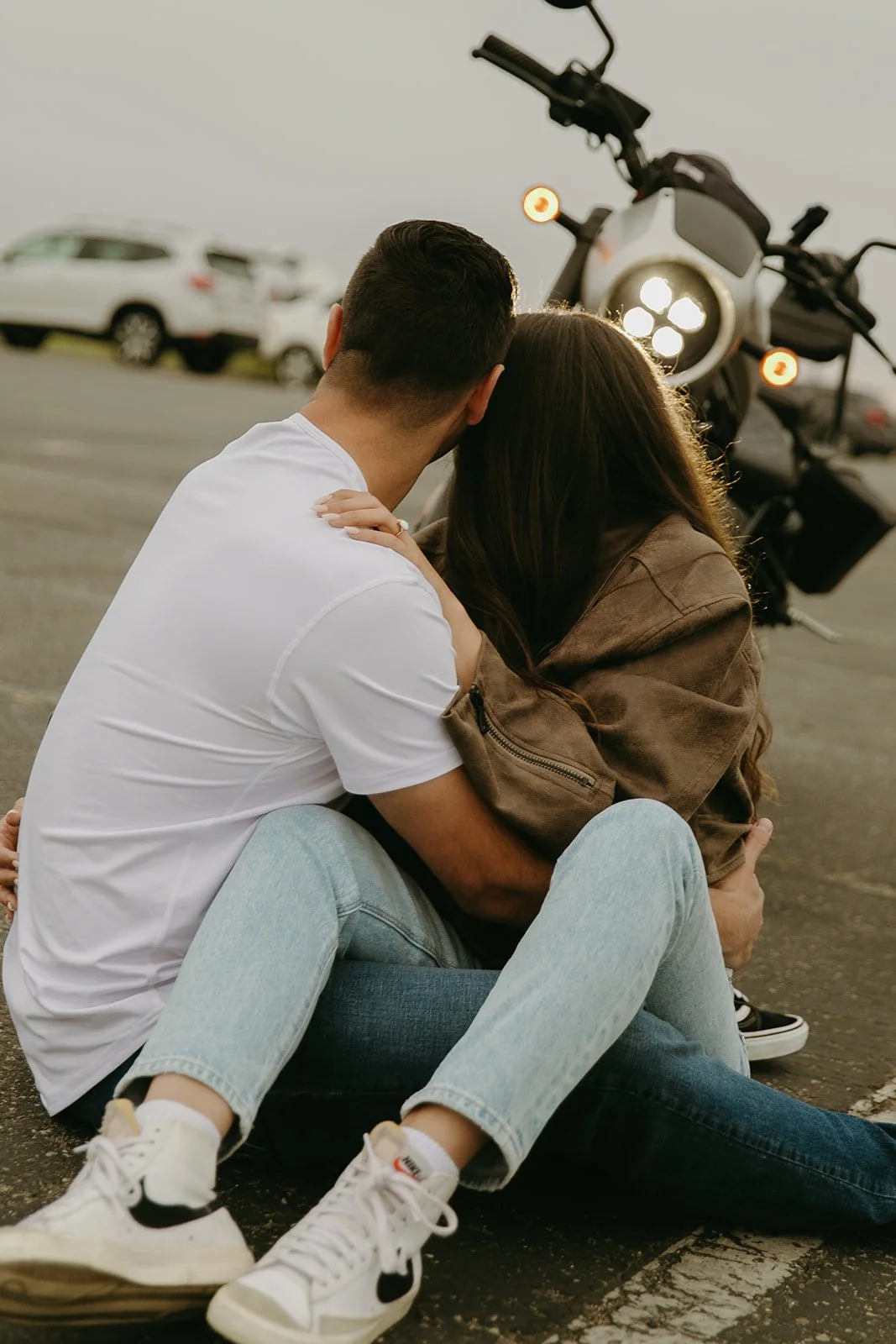 A couple sits close together on the pavement, leaning in with a motorcycle glowing behind them.