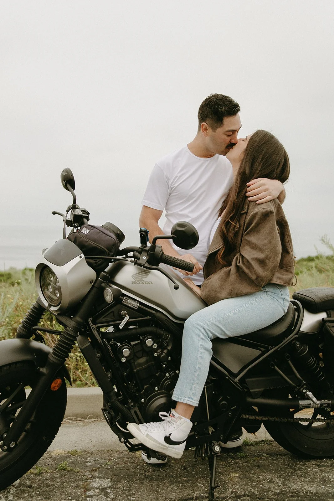 A couple shares a soft kiss while seated on a motorcycle, surrounded by greenery — a perfect blend of adventure and romantic engagement photo ideas.