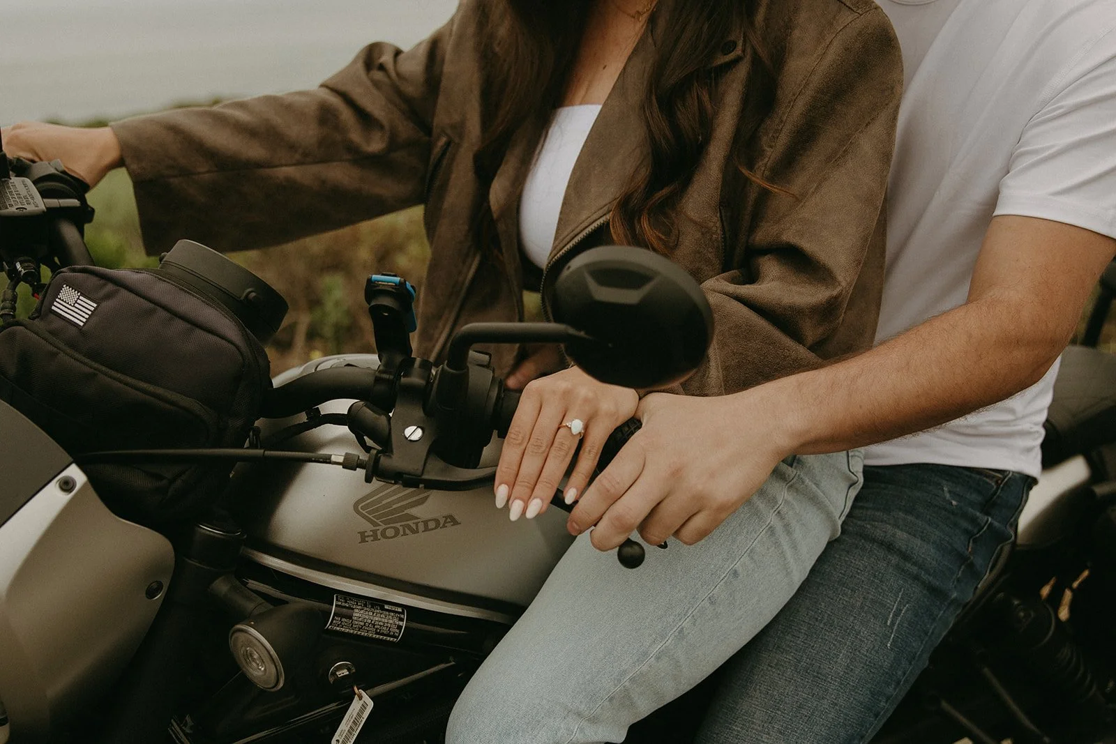 Close-up of a couple’s hands resting on a motorcycle handlebar, showing off an engagement ring and cozy embrace.