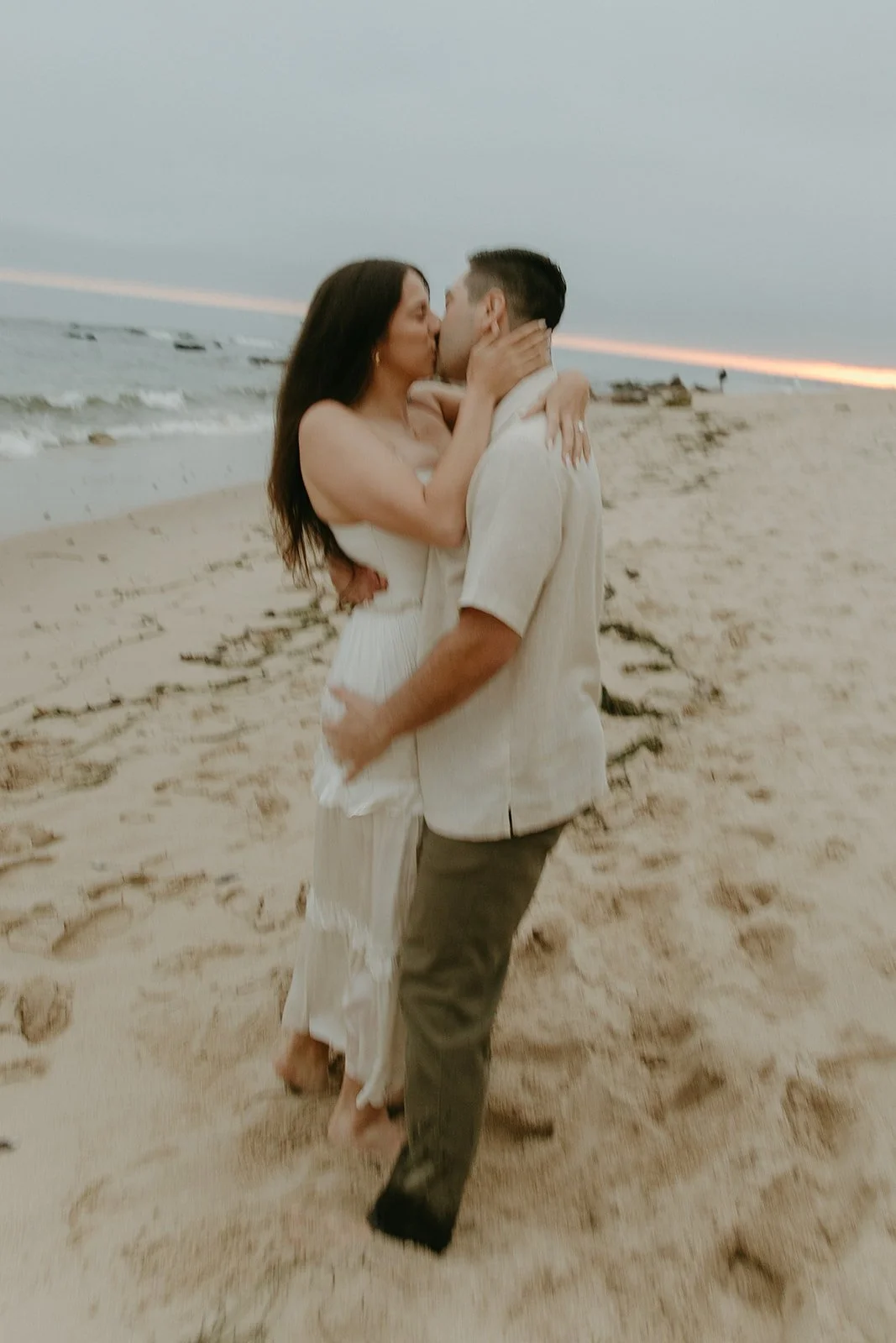 A soft, slightly blurred photo of a couple kissing on the beach at sunset, standing barefoot in the sand.