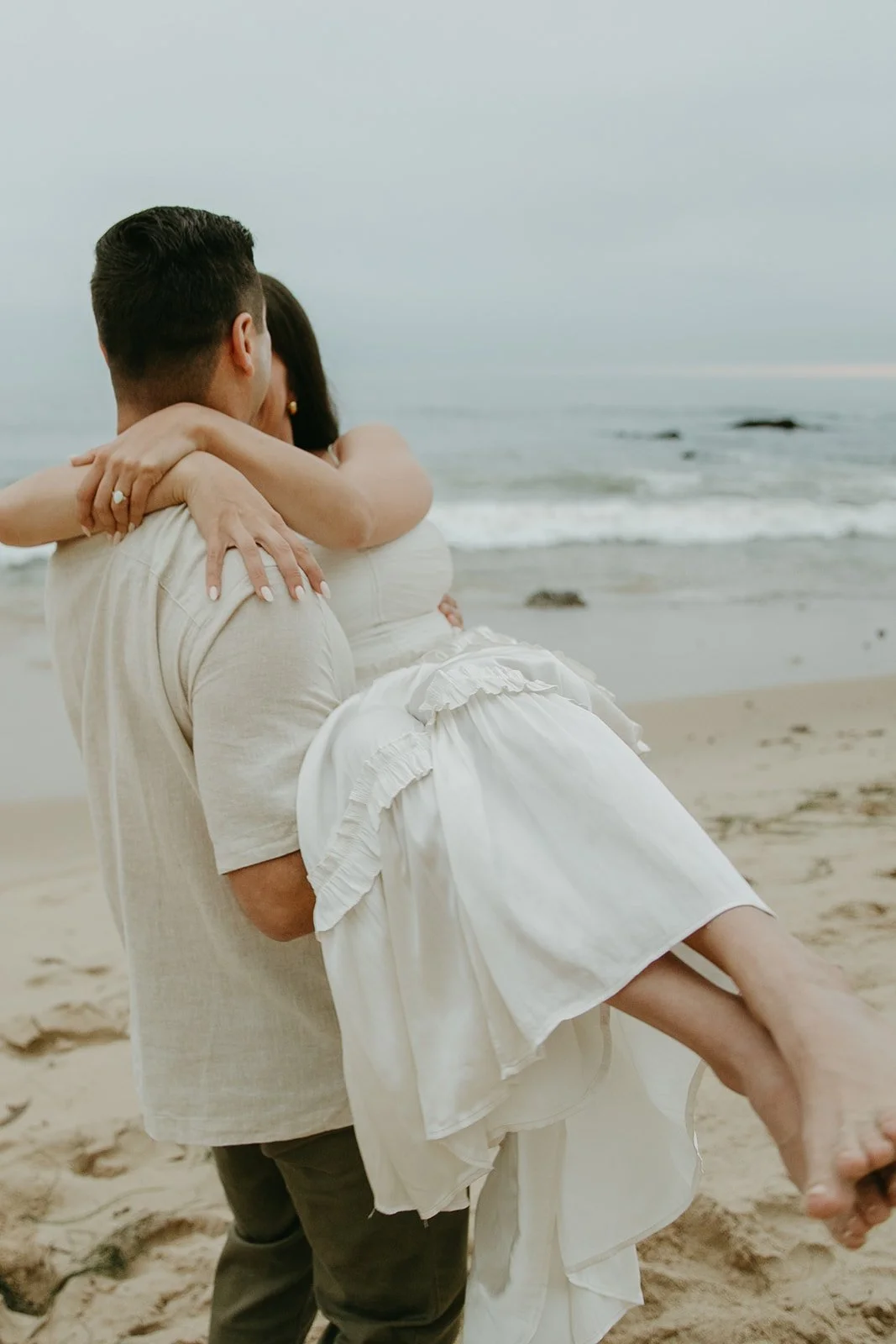 A groom-to-be carries his partner on the beach as they share an intimate moment by the ocean.