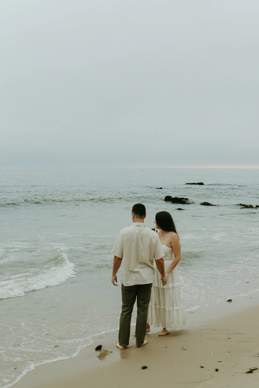 A couple on the beach holding hands and watching the waves crash as the sun sets. A beautiful way to document romantic engagement photo ideas.