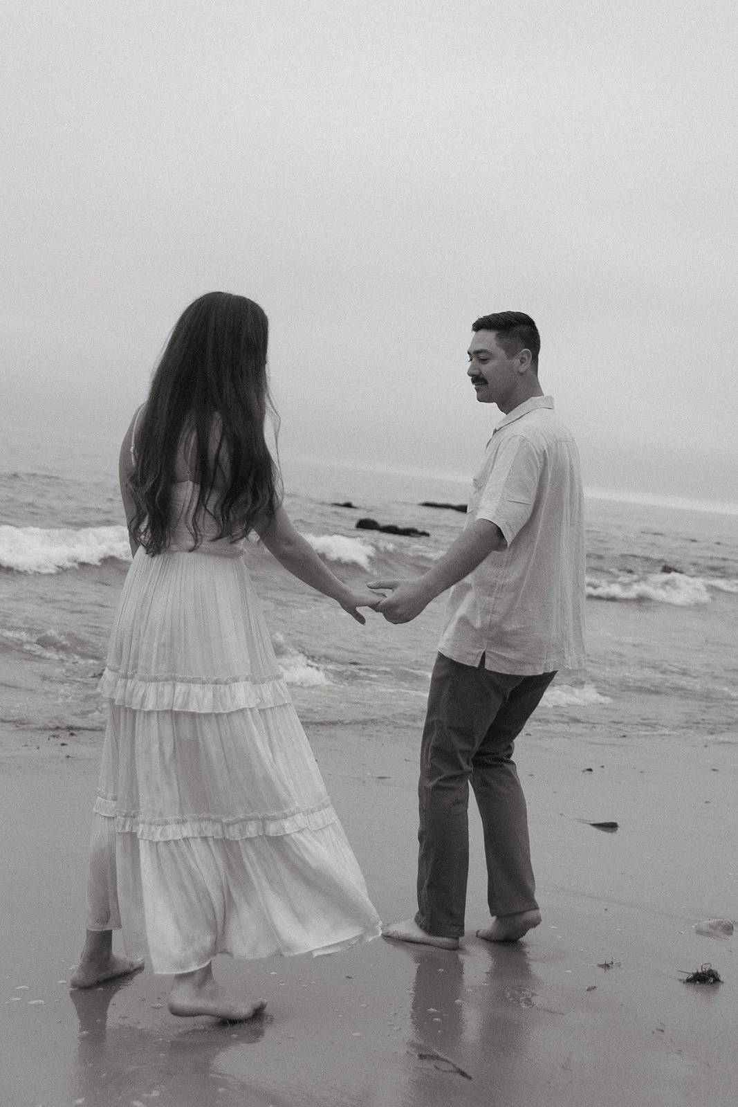 A black and white photo of a couple walking barefoot on the beach, holding hands with gentle waves nearby, a dreamy concept for romantic engagement photo ideas.