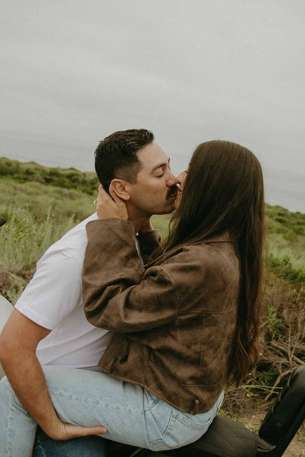 A couple kisses while sitting on a motorcycle, surrounded by coastal greenery.