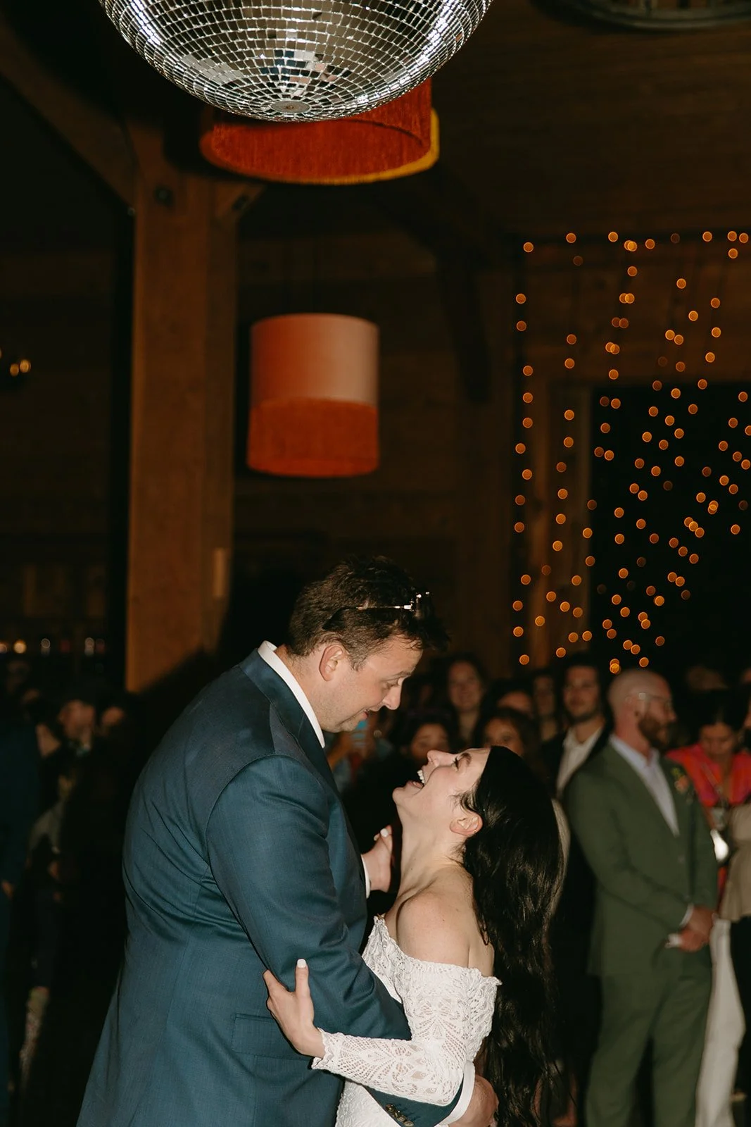 The bride and groom share their first dance beneath a disco ball, surrounded by guests and twinkle lights.