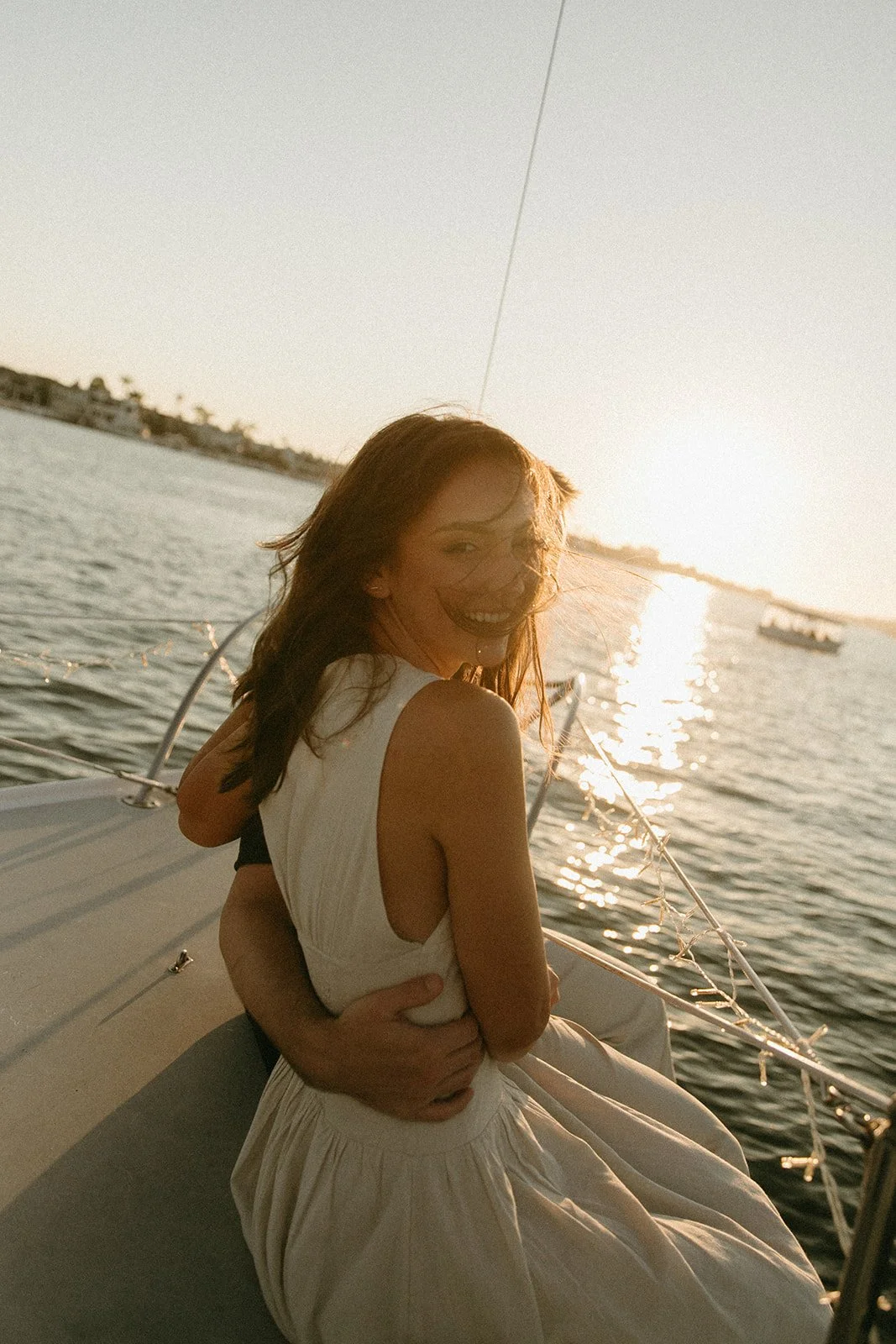 Woman smiling at sunset while being held by her partner on a sailboat, sunset sailboat engagement photos with golden hour light.