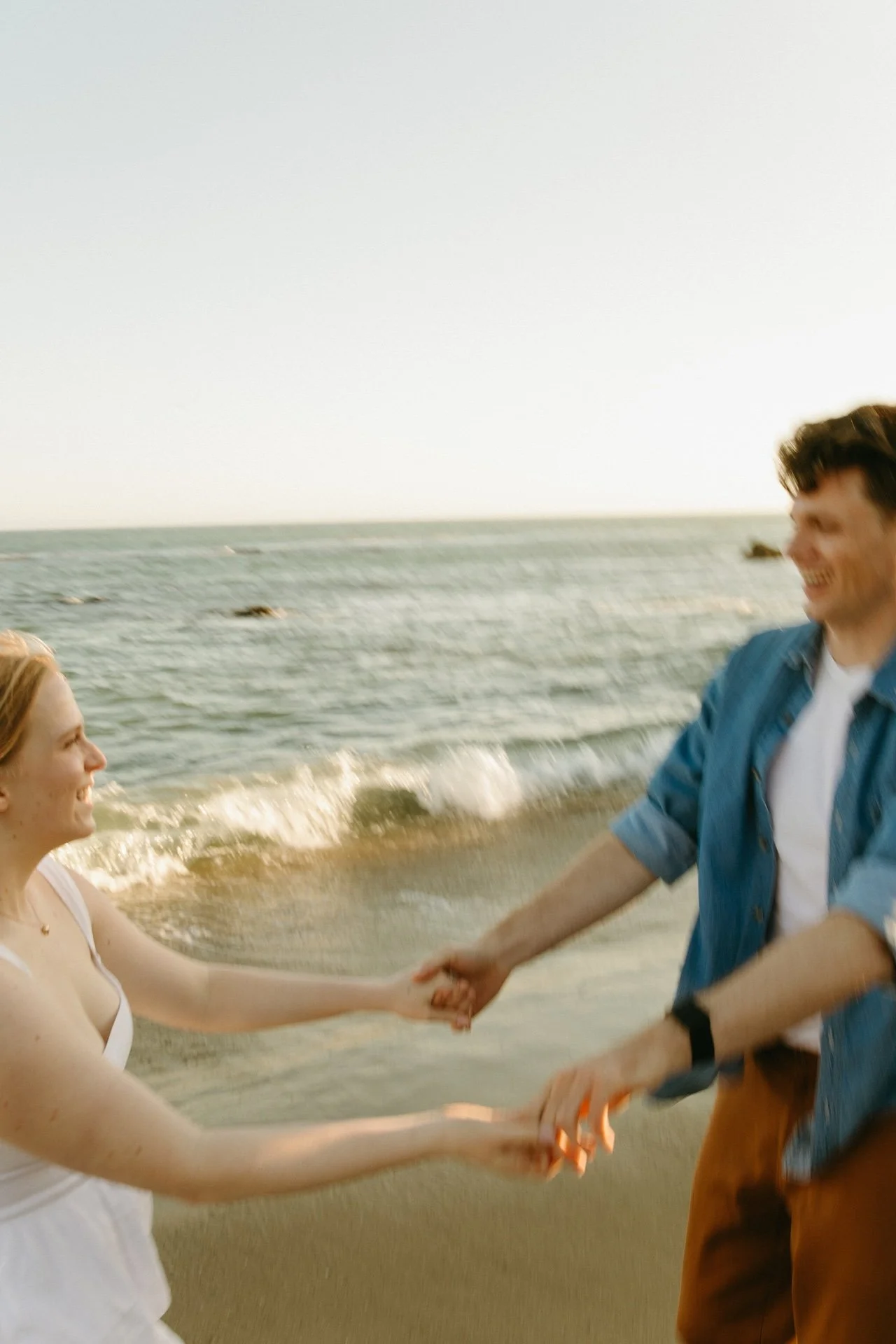 Couple holding hands and playfully spinning near the waterline, with the beach and waves creating a joyful scene at one of the most fun engagement photo locations in Malibu.