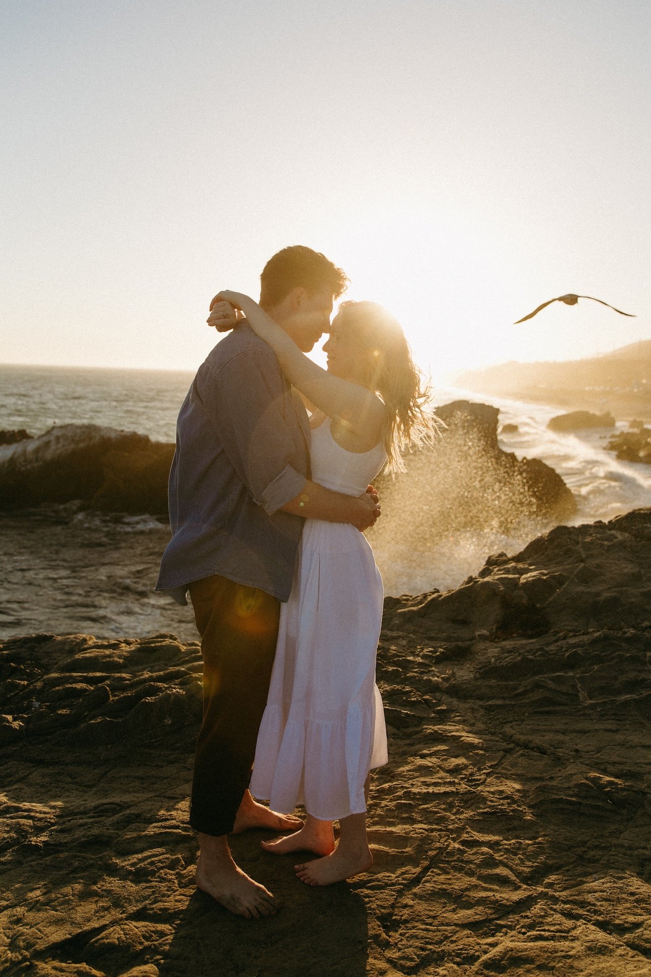 Couple embracing on rocky cliffs as ocean waves crash behind them, golden sunlight streaming through at one of the most breathtaking engagement photo locations.