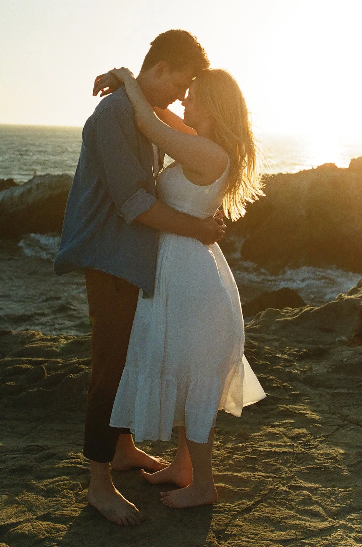 Couple hugging and touching forheads on a rocky cliff on the ocean with the sun setting behind them during their Malibu engagement shoot.