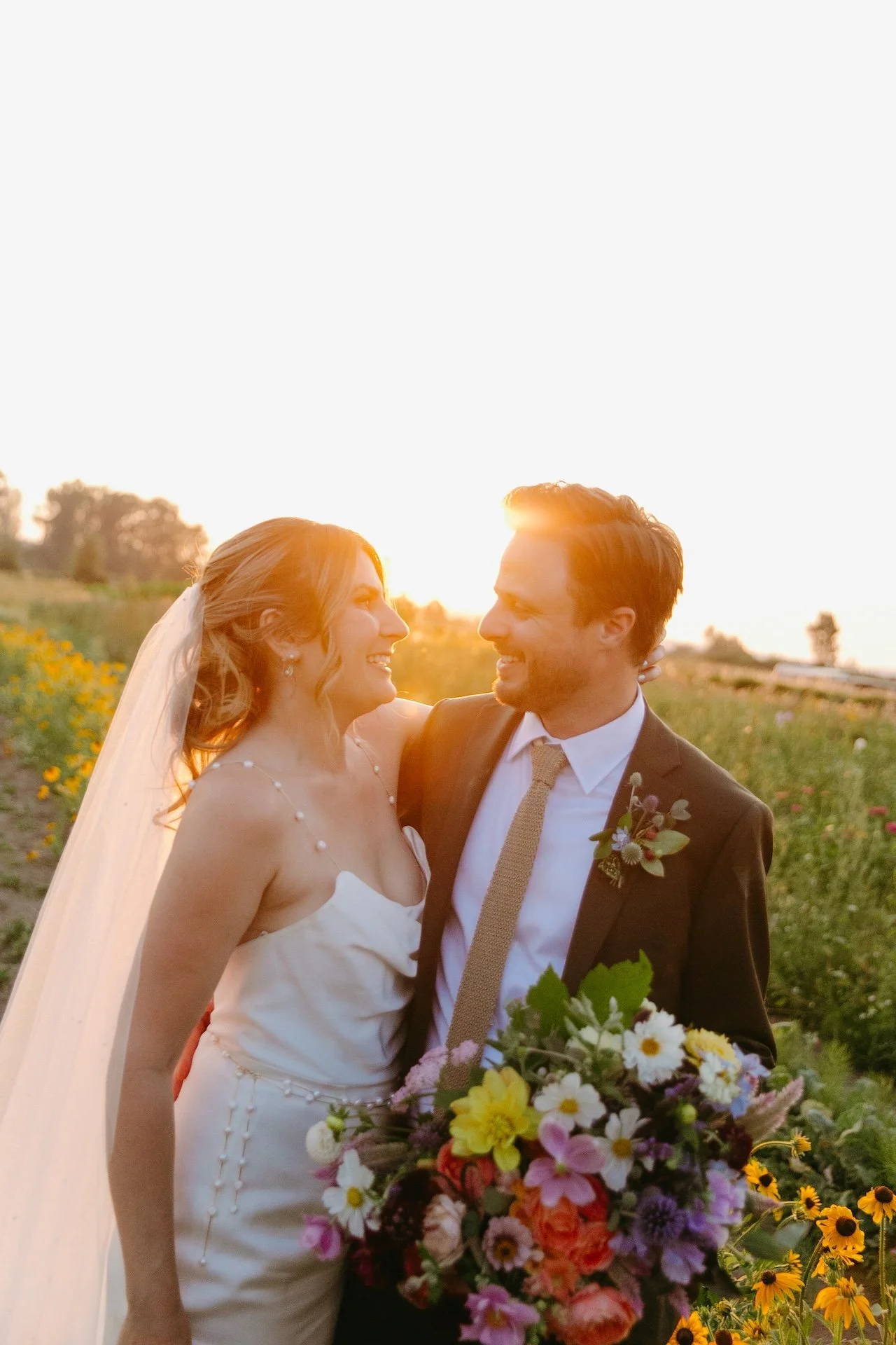 Smiling bride and groom embrace in a field of wildflowers at sunset, celebrating their garden party wedding.