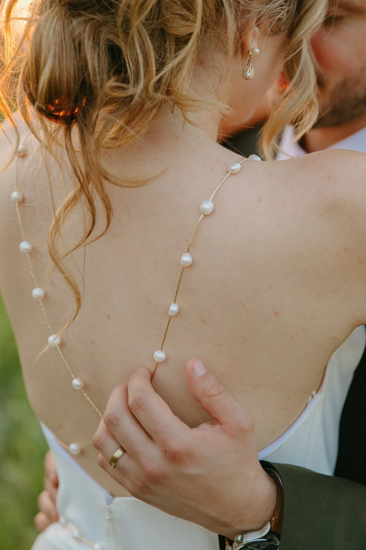 Close-up of the bride’s pearl-detailed gown strap and groom’s hand on her back.