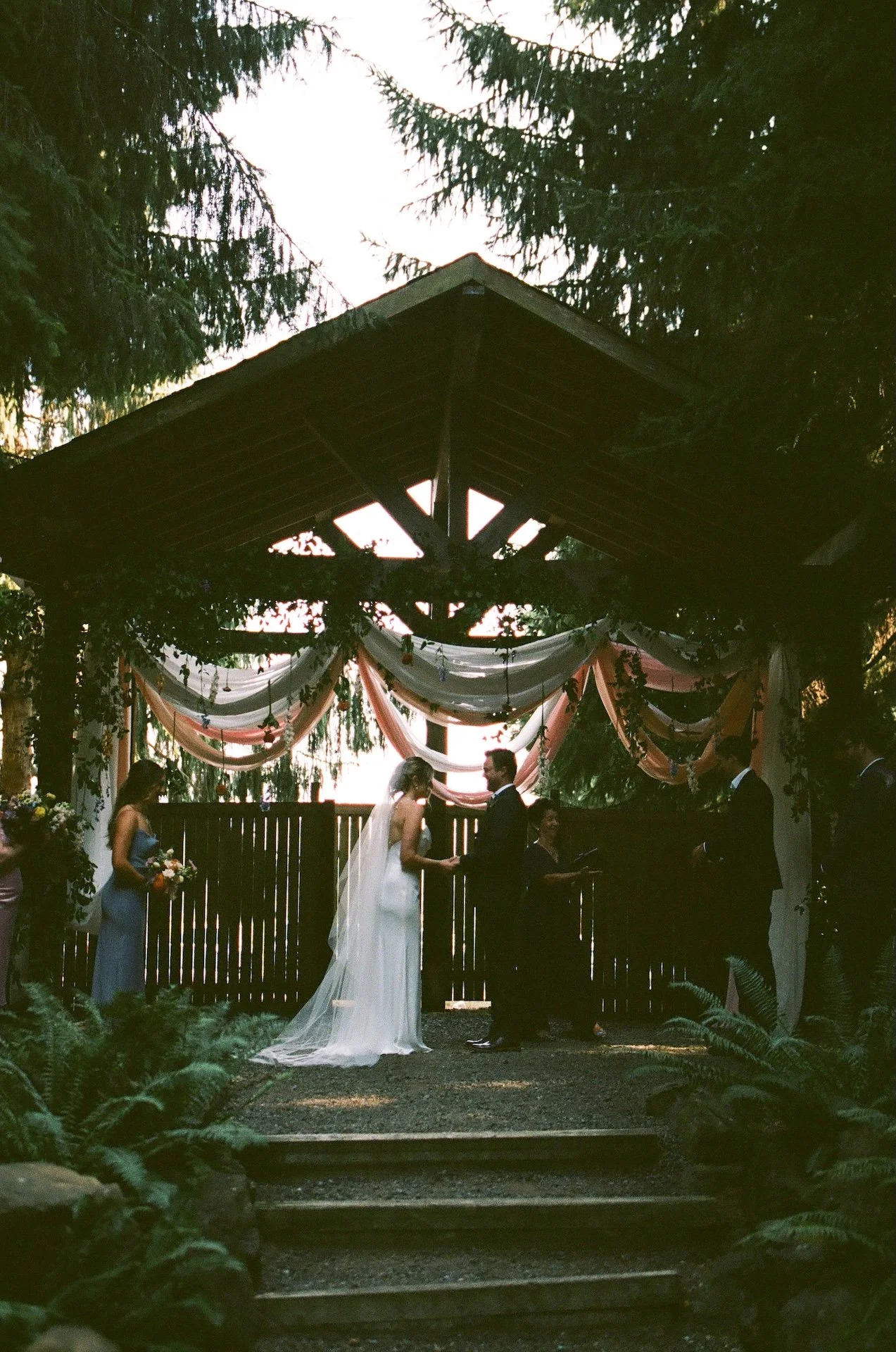 Bride and groom stand beneath the arbor exchanging vows, with bridesmaids and groomsmen on either side at their garden party wedding.