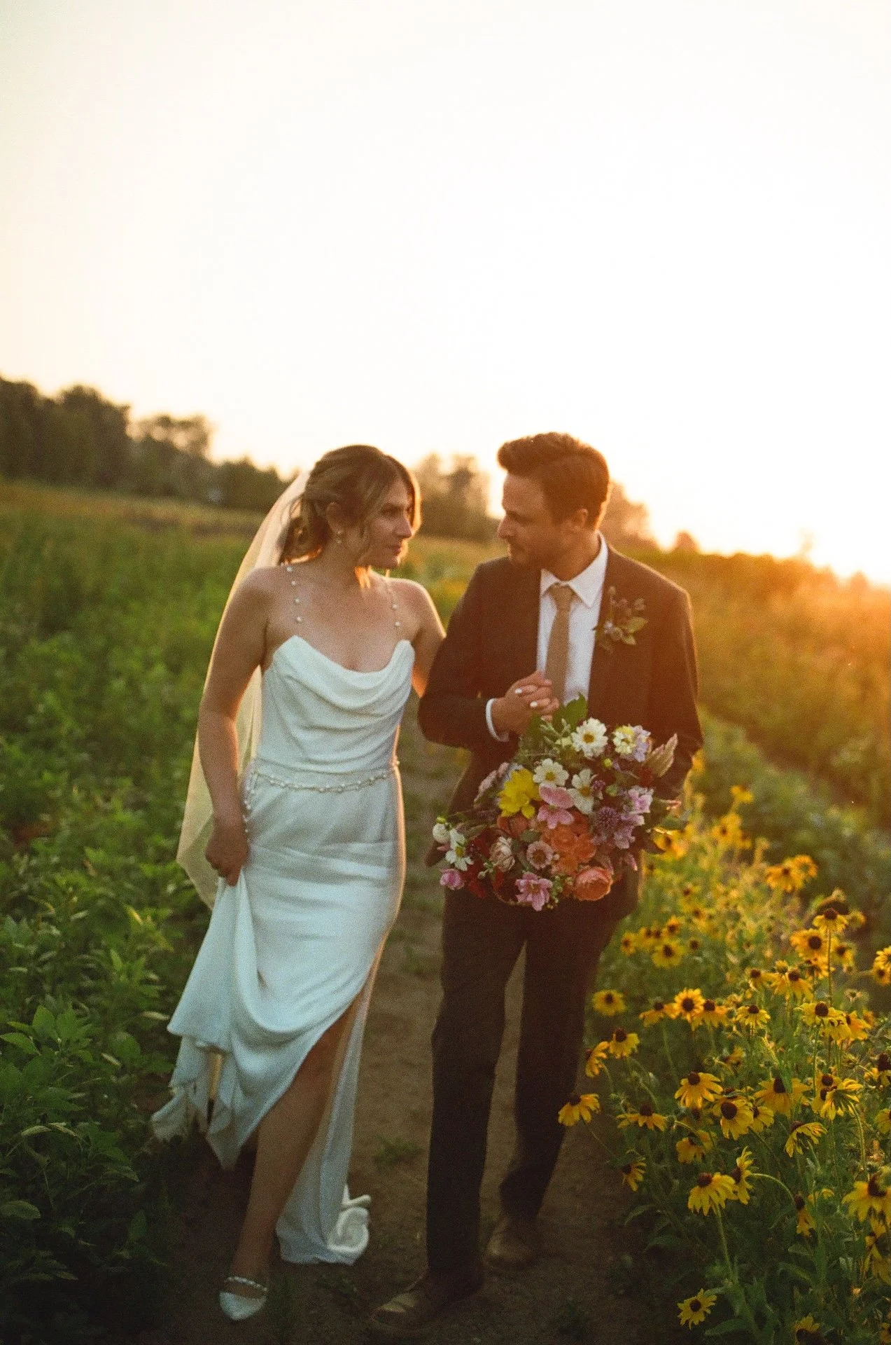 Newlyweds walk through a field of flowers at sunset, holding hands and a bouquet at their garden party wedding.