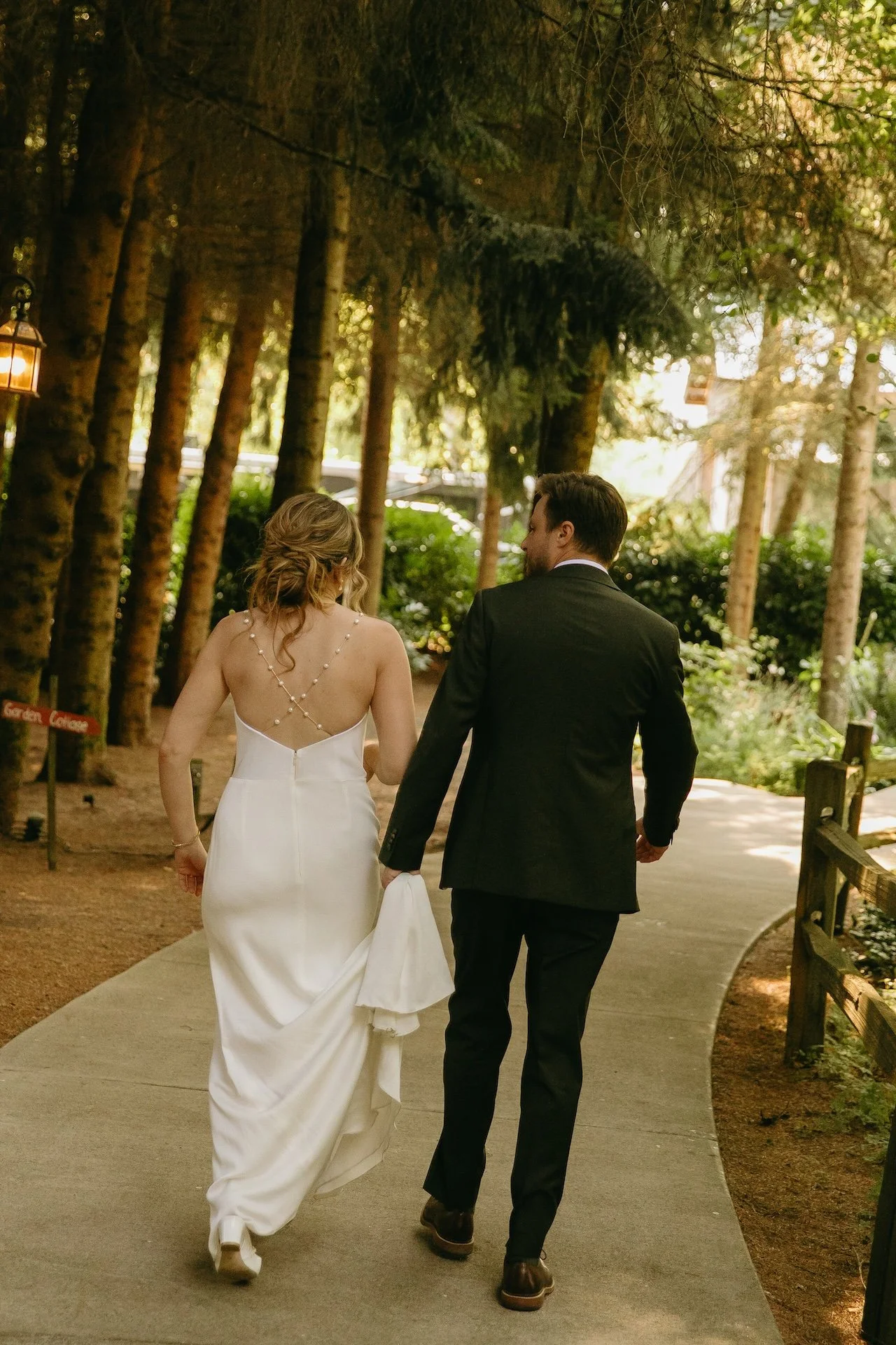 Couple walks down a forest path hand in hand, the bride’s dress flowing behind her.