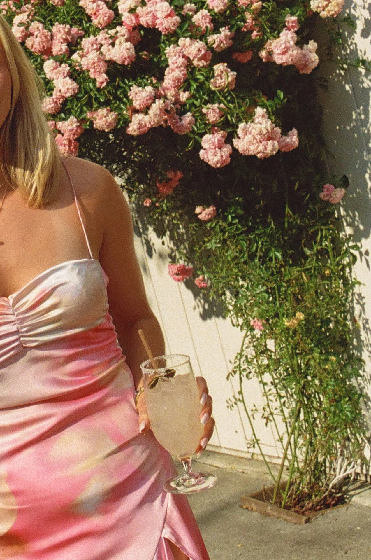 Guest in a pink dress holds a refreshing cocktail in front of a wall of blooming pink roses.