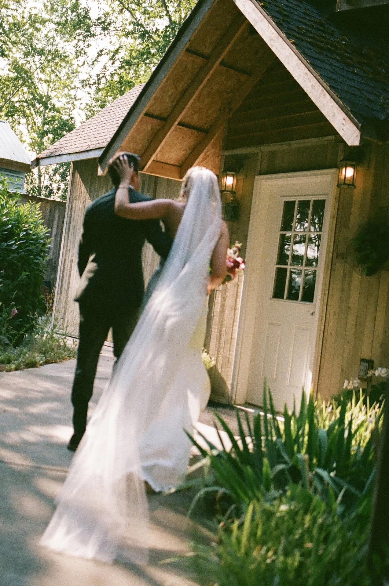 Bride and groom walk arm in arm toward a rustic wooden building at their garden party wedding venue.